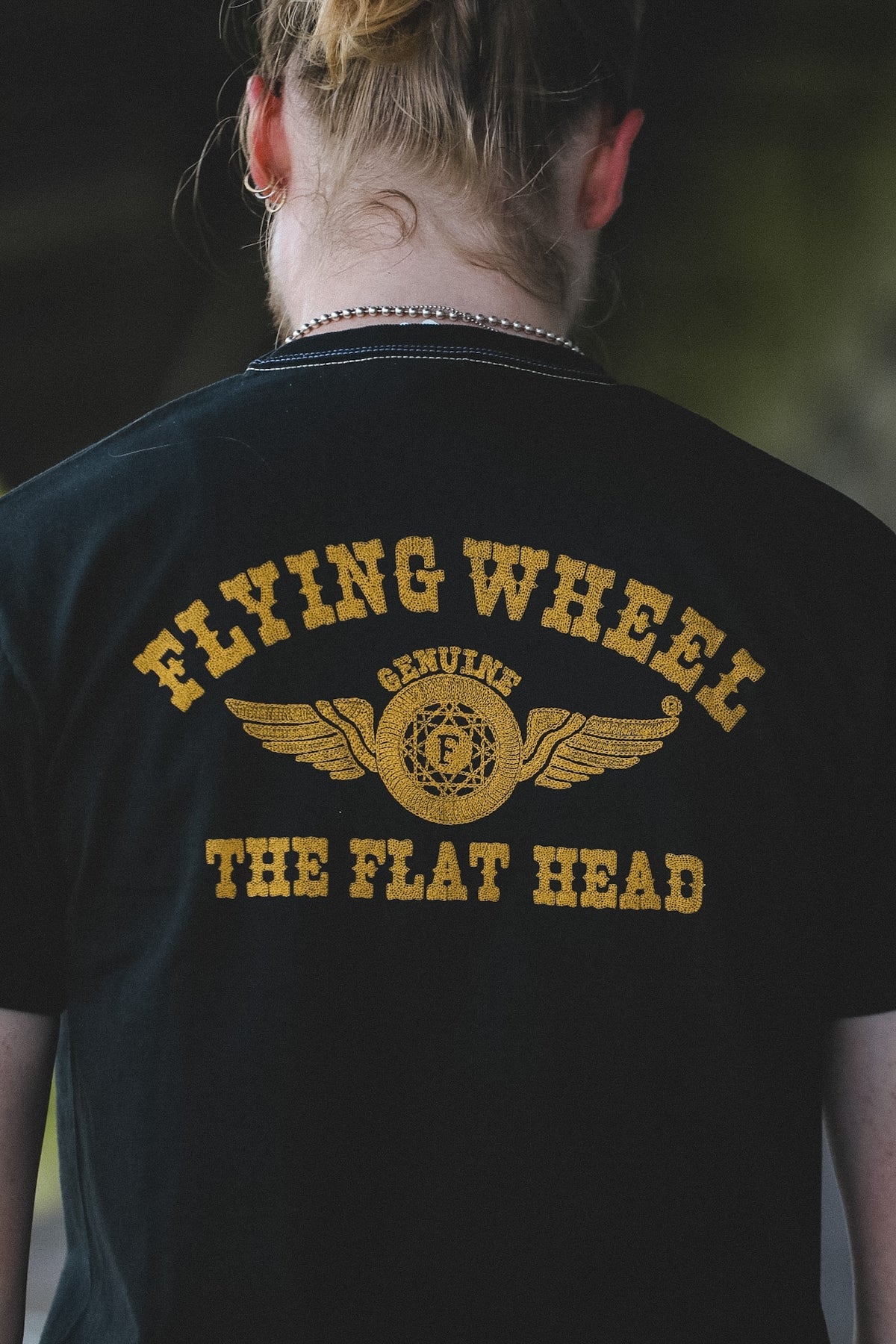A person viewed from behind wearing a black t-shirt with a yellow graphic that reads 'FLYING WHEEL' and 'THE FLAT HEAD' with an airplane wing emblem. The person is walking along an outdoor covered walkway next to a building.
