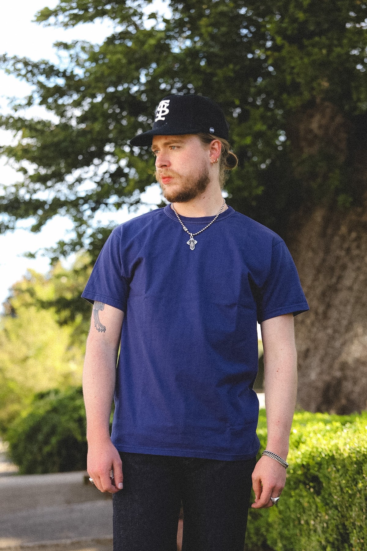 A man wearing a navy blue crewneck t-shirt, a black baseball cap with a white logo, and a silver chain necklace, standing outdoors in front of a large tree.