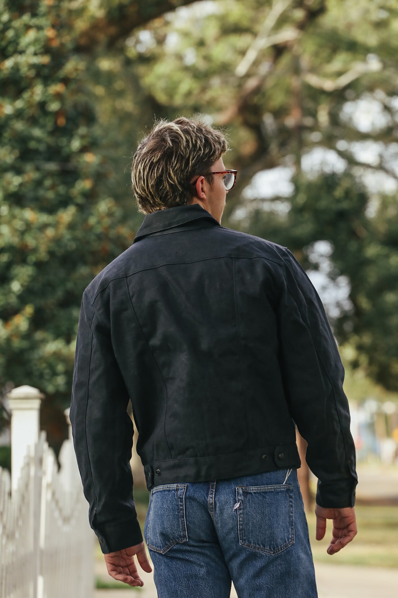 Man wearing a black jacket and blue jeans walking away from the camera with trees in the background