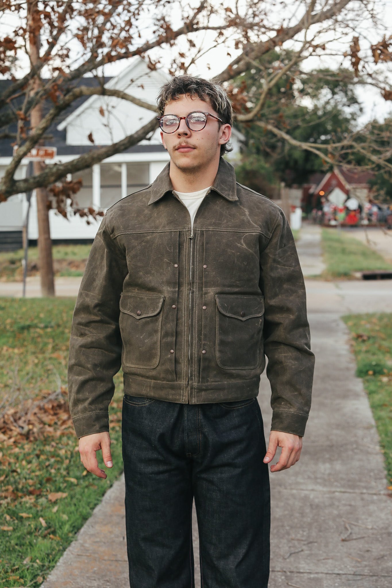 Man wearing a brown jacket and glasses standing on a sidewalk with trees and houses in the background.