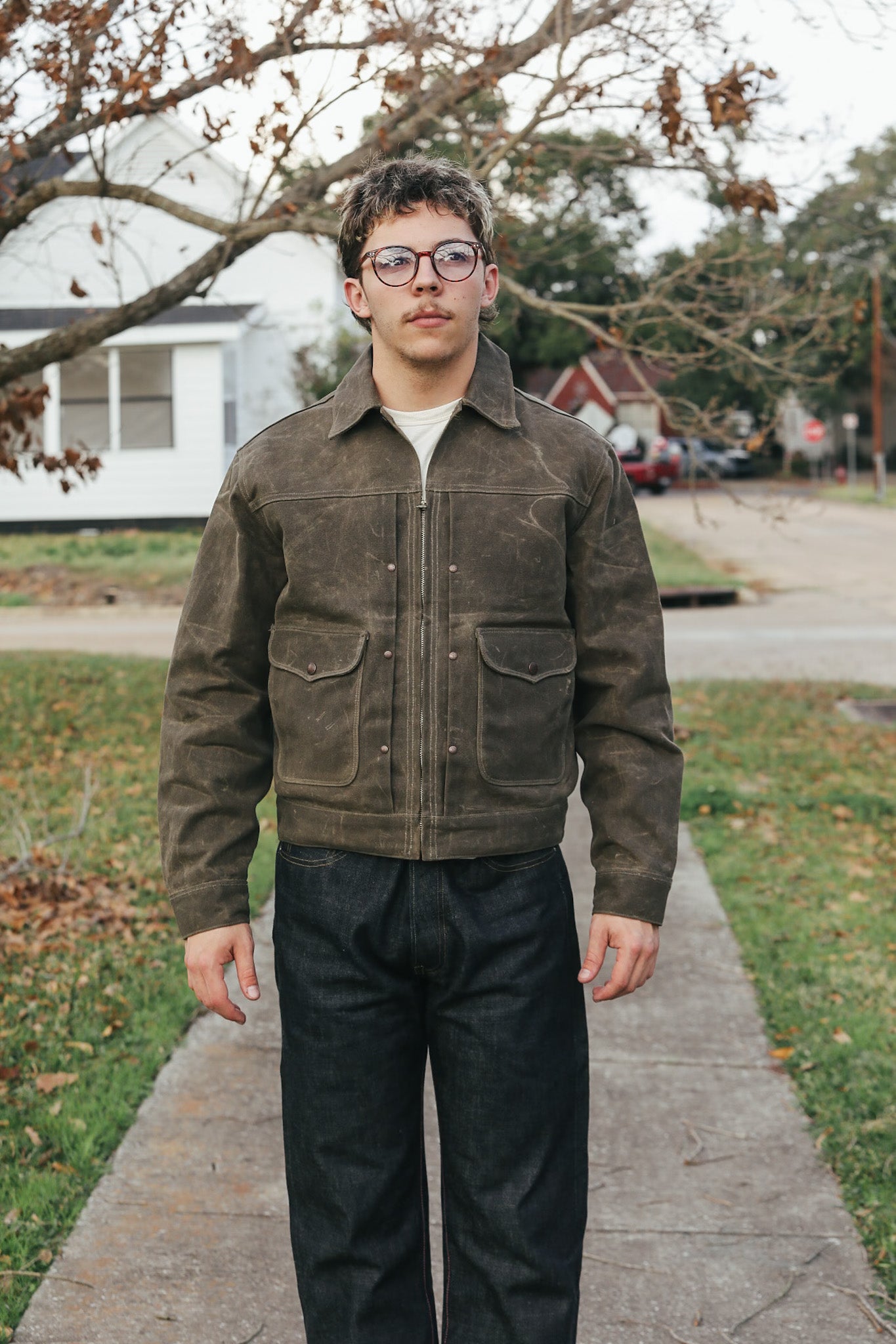 Man wearing a brown jacket and black pants standing on a sidewalk with a residential background.