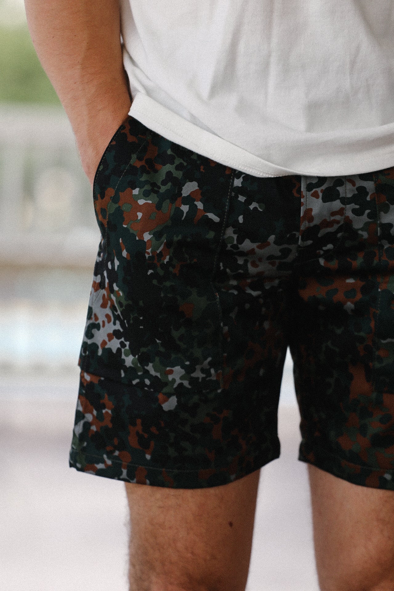 Close-up of a person wearing diffusion camo fatigue shorts and a white t-shirt, hands in pockets