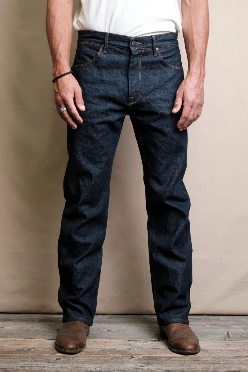 Man wearing dark blue raw denim jeans and brown leather boots standing on wooden floor against beige backdrop