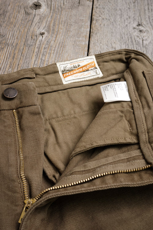 Close-up of olive green Freenote Cloth pants with zipper and label on rustic wooden surface
