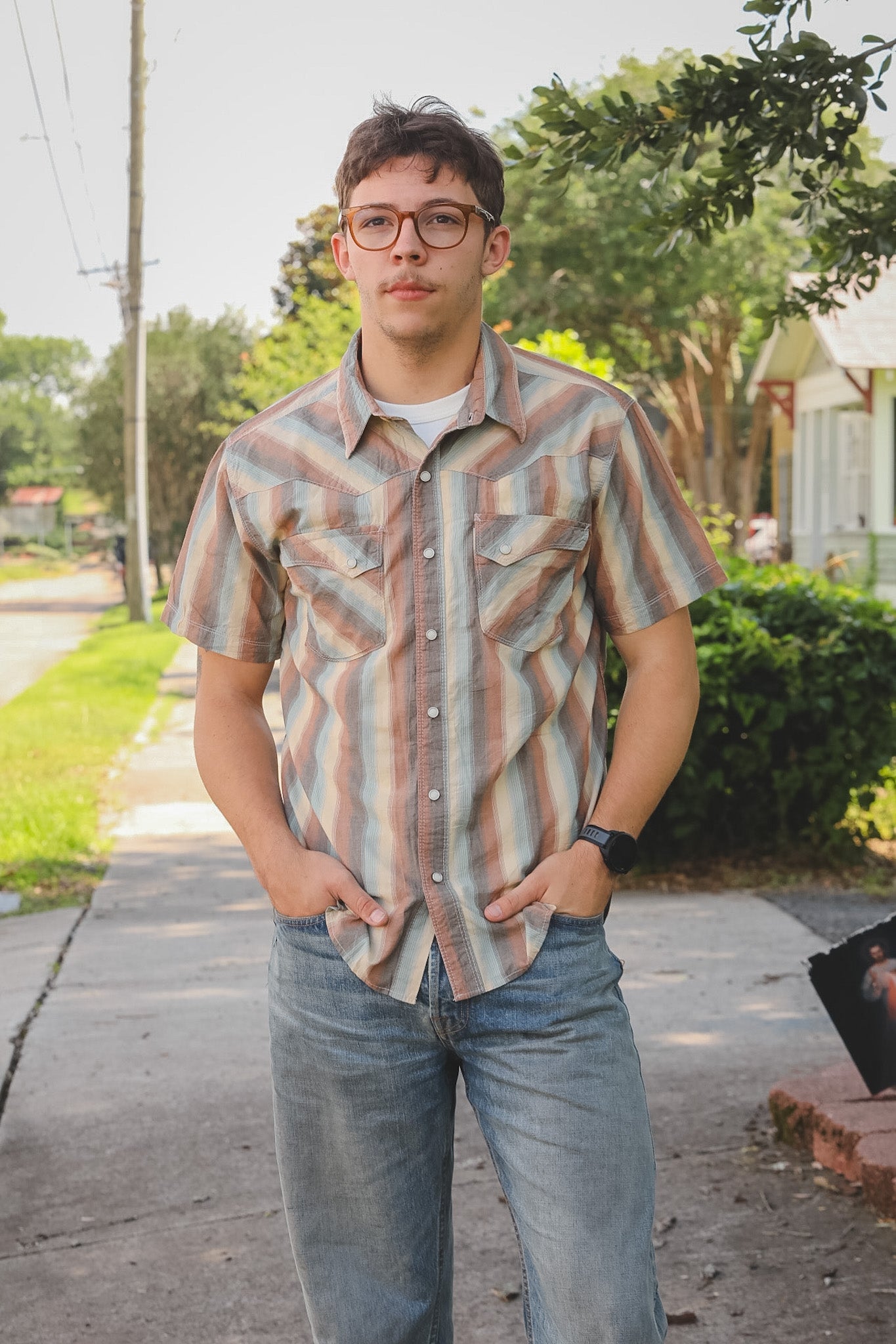Man wearing a patterned shirt and jeans standing outdoors on a sidewalk.