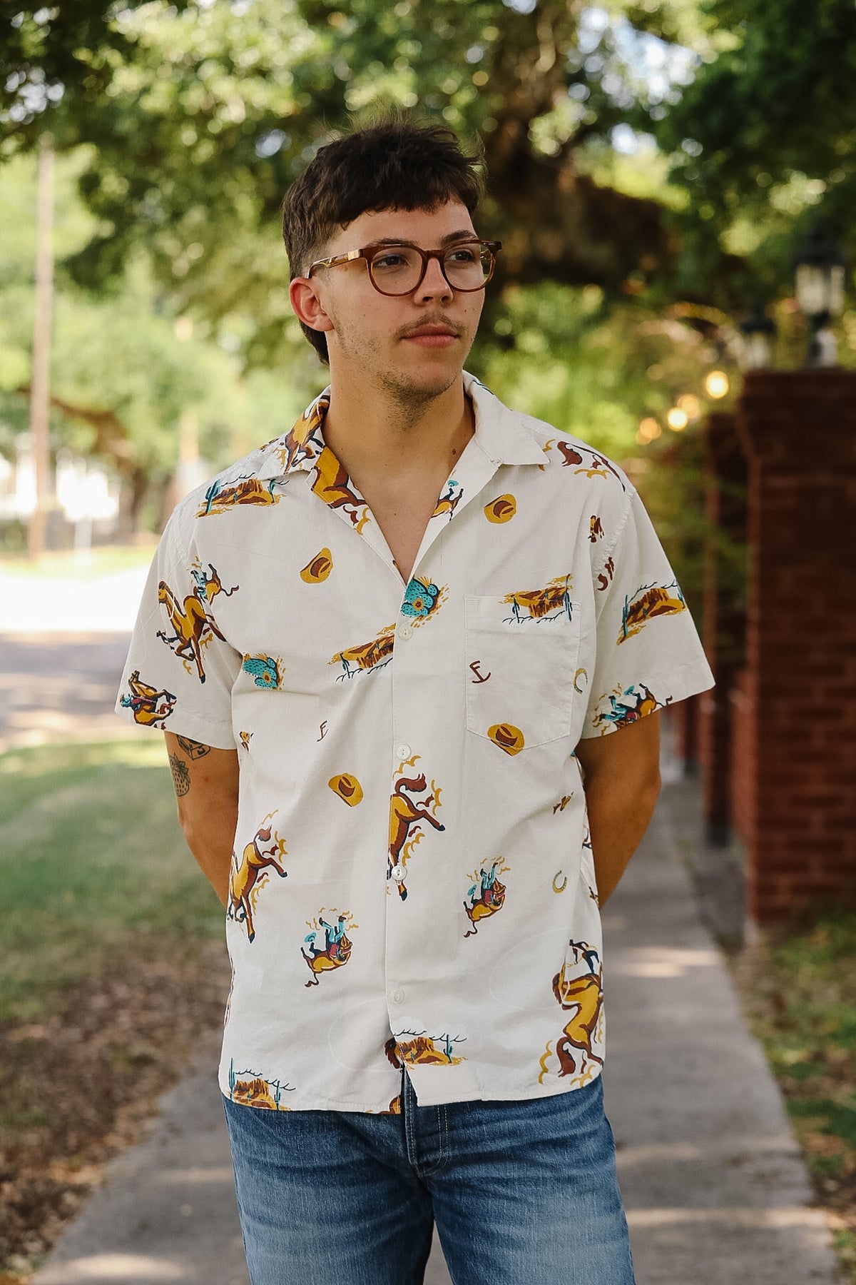 A man wearing a white short-sleeve button-up shirt with a colorful cowboy and horseshoe pattern, paired with blue jeans, standing outdoors on a sidewalk with trees in the background.