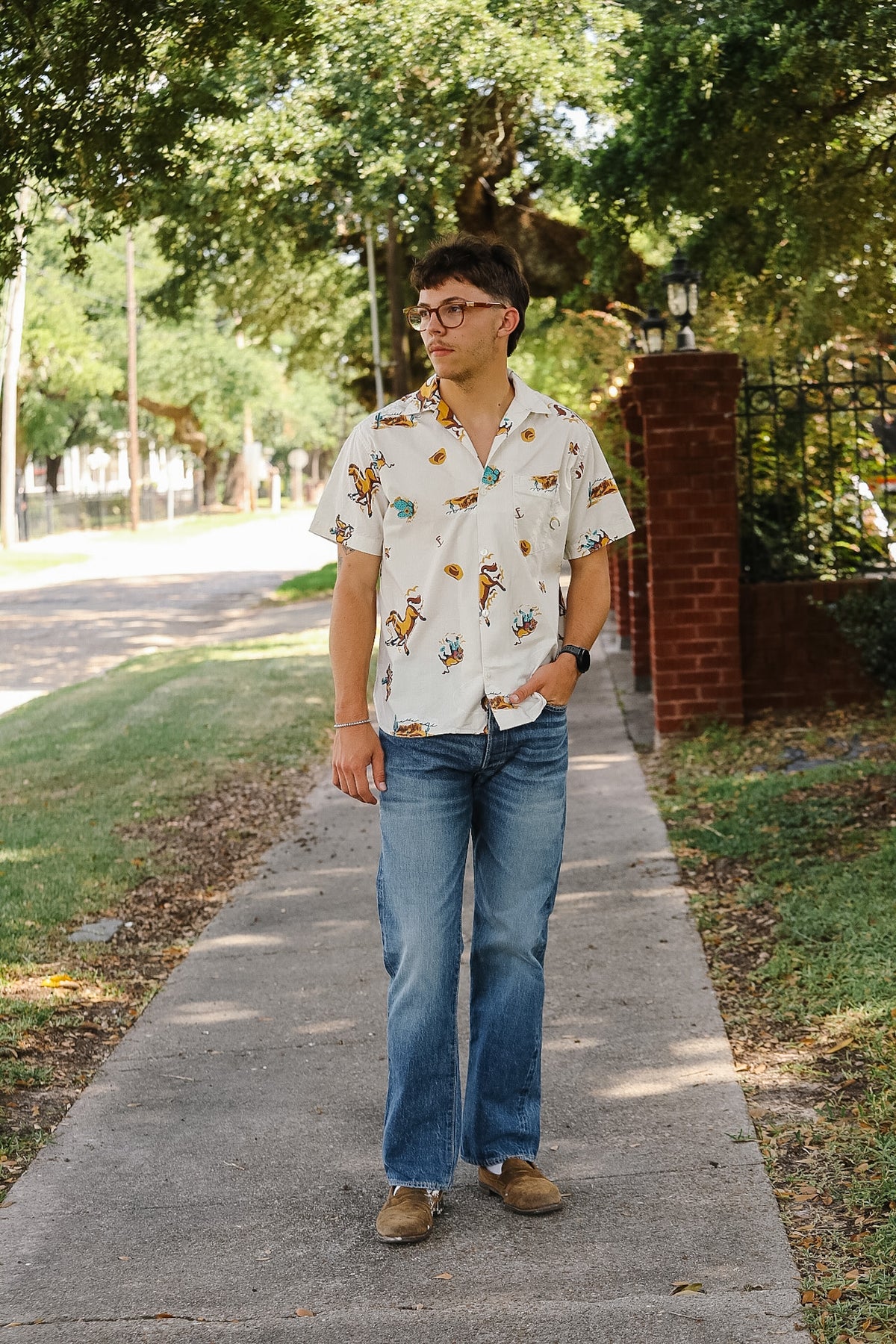 A man wearing a white short-sleeve button-down shirt with a colorful cartoon animal print, blue jeans, and brown shoes, standing on a paved sidewalk in a tree-lined street.