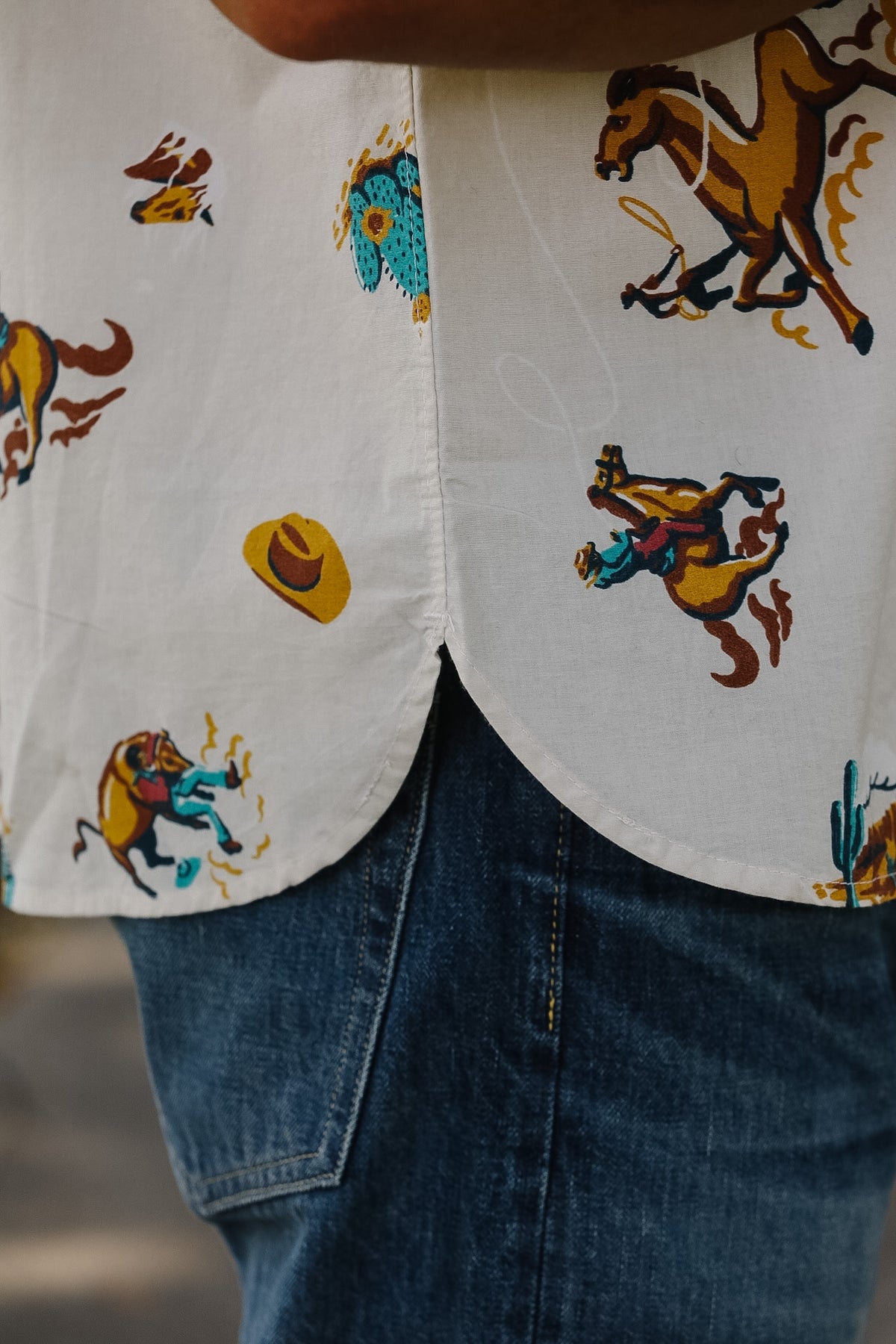 Close-up of a person wearing a white button-up shirt with a colorful western print of horses, riders, and hats, paired with blue jeans.