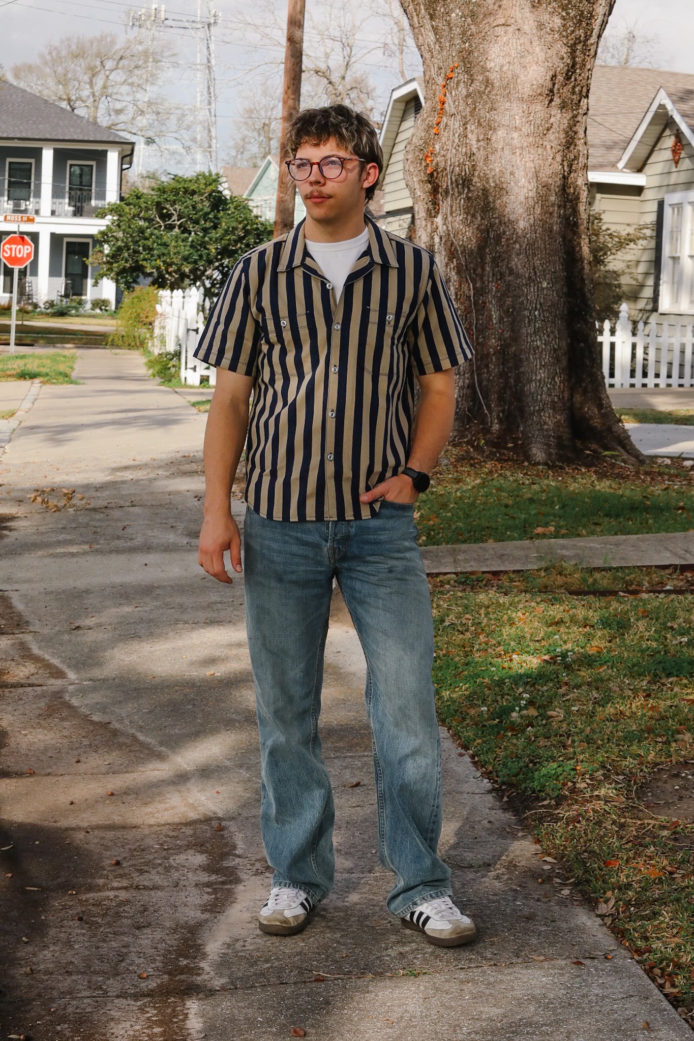 Person wearing a striped shirt and jeans standing on a sidewalk with houses and trees in the background.