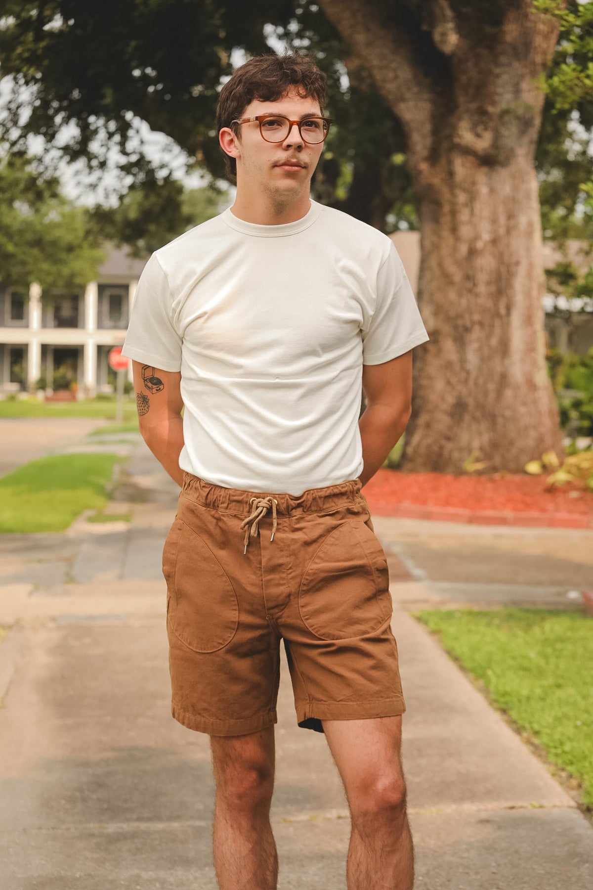 Man wearing a white t-shirt and brown shorts standing on a sidewalk with trees and a house in the background.