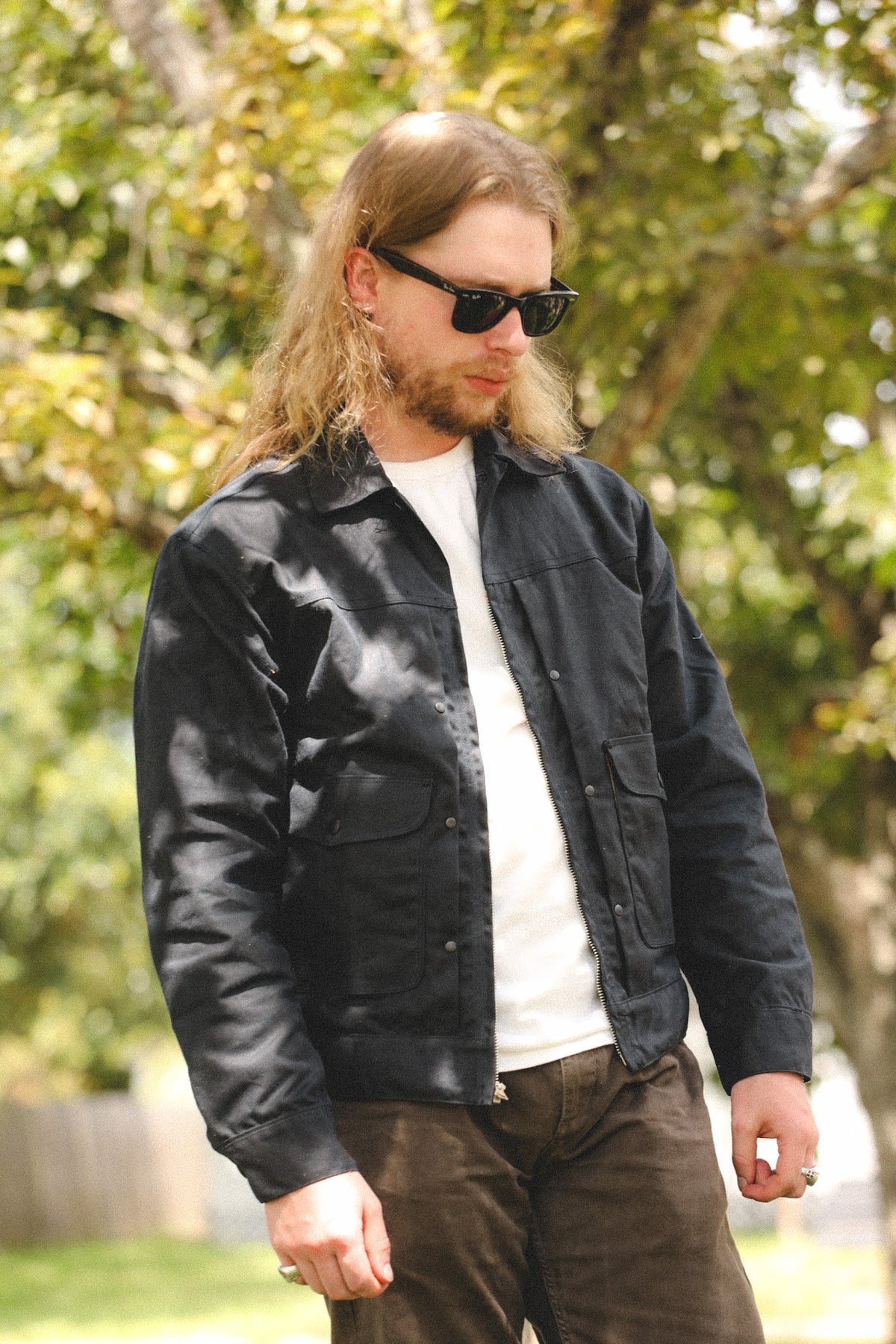 A man with long blonde hair and sunglasses wearing a black overshirt over a white t-shirt and brown pants, standing outdoors in front of a blurred background of trees.