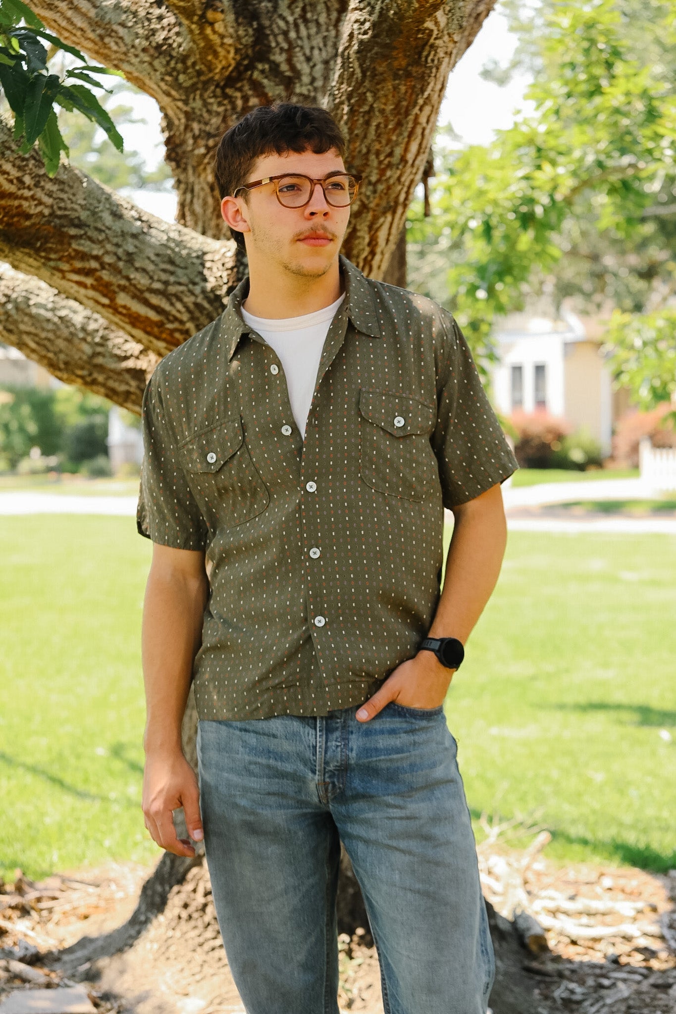 Person wearing a patterned shirt and jeans standing outdoors near a tree