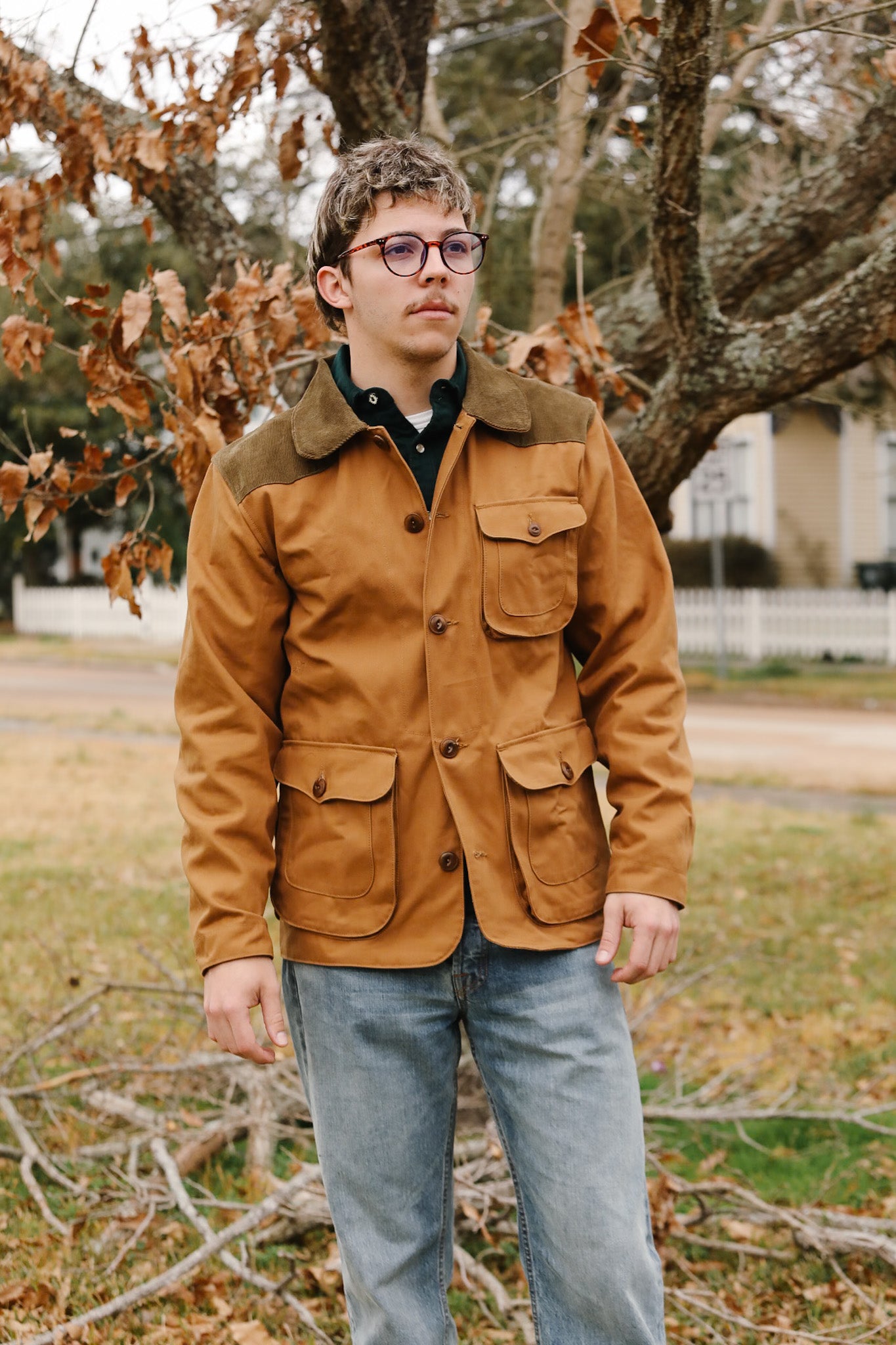 Man wearing a brown jacket with fur collar standing outdoors near trees and grass.