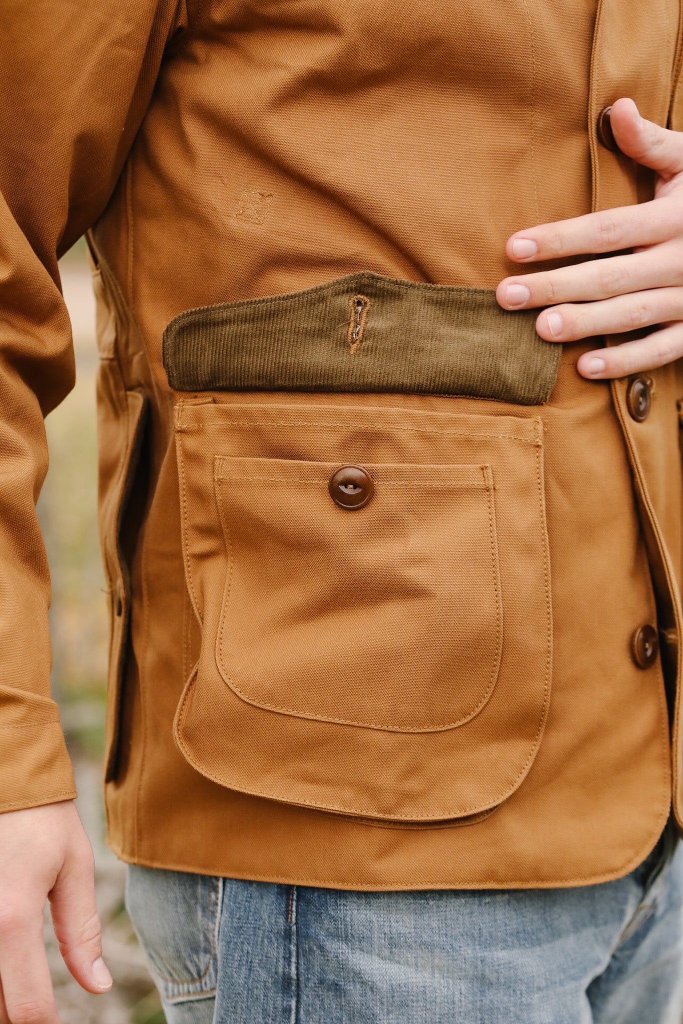 Close-up of a brown jacket with a pocket and button, worn over jeans.