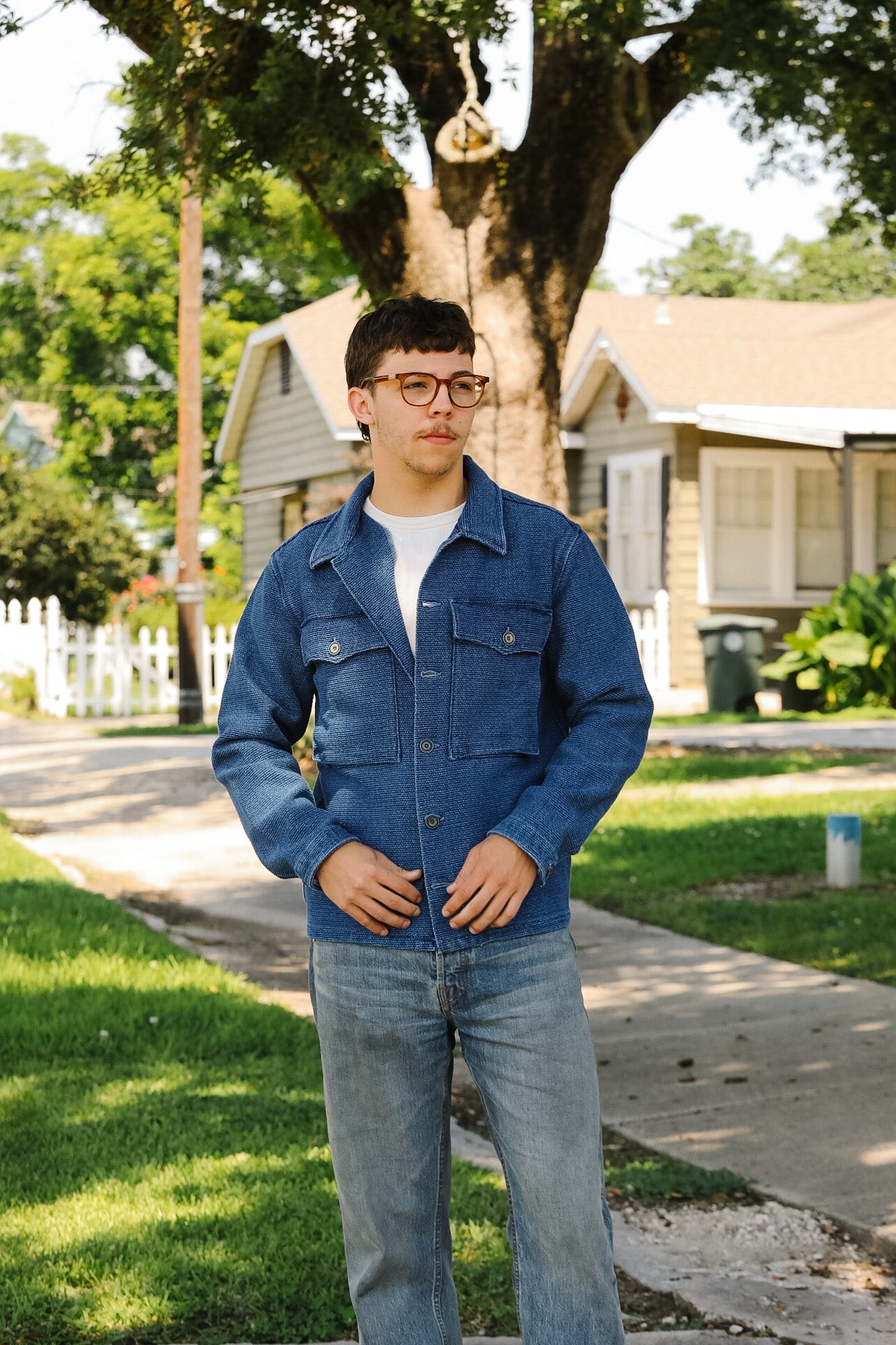 Person wearing a blue jacket and jeans standing on a sidewalk with houses and trees in the background