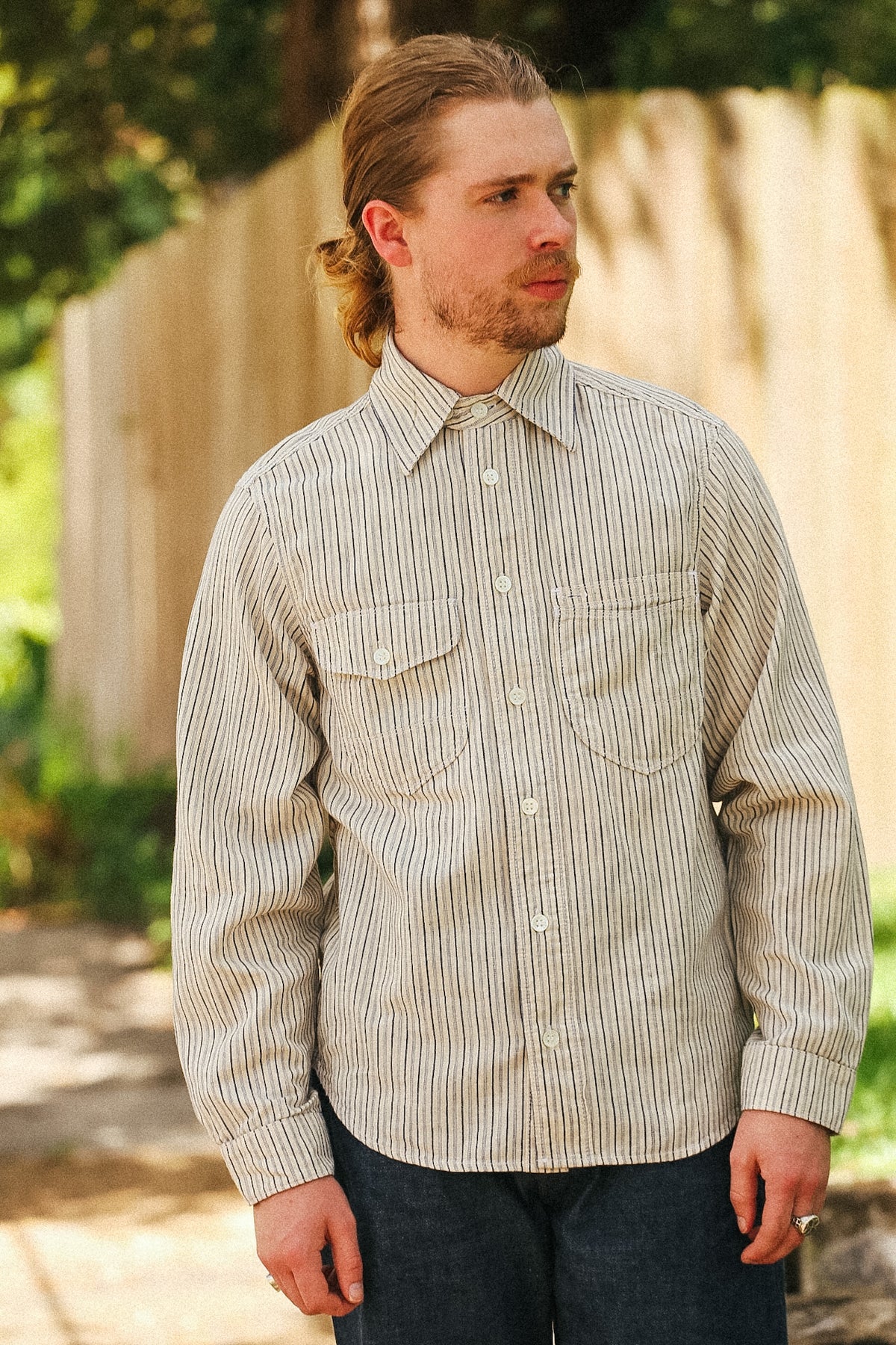 A man wearing a beige and white vertically striped long-sleeve button-down shirt, standing outdoors in front of a wooden fence.