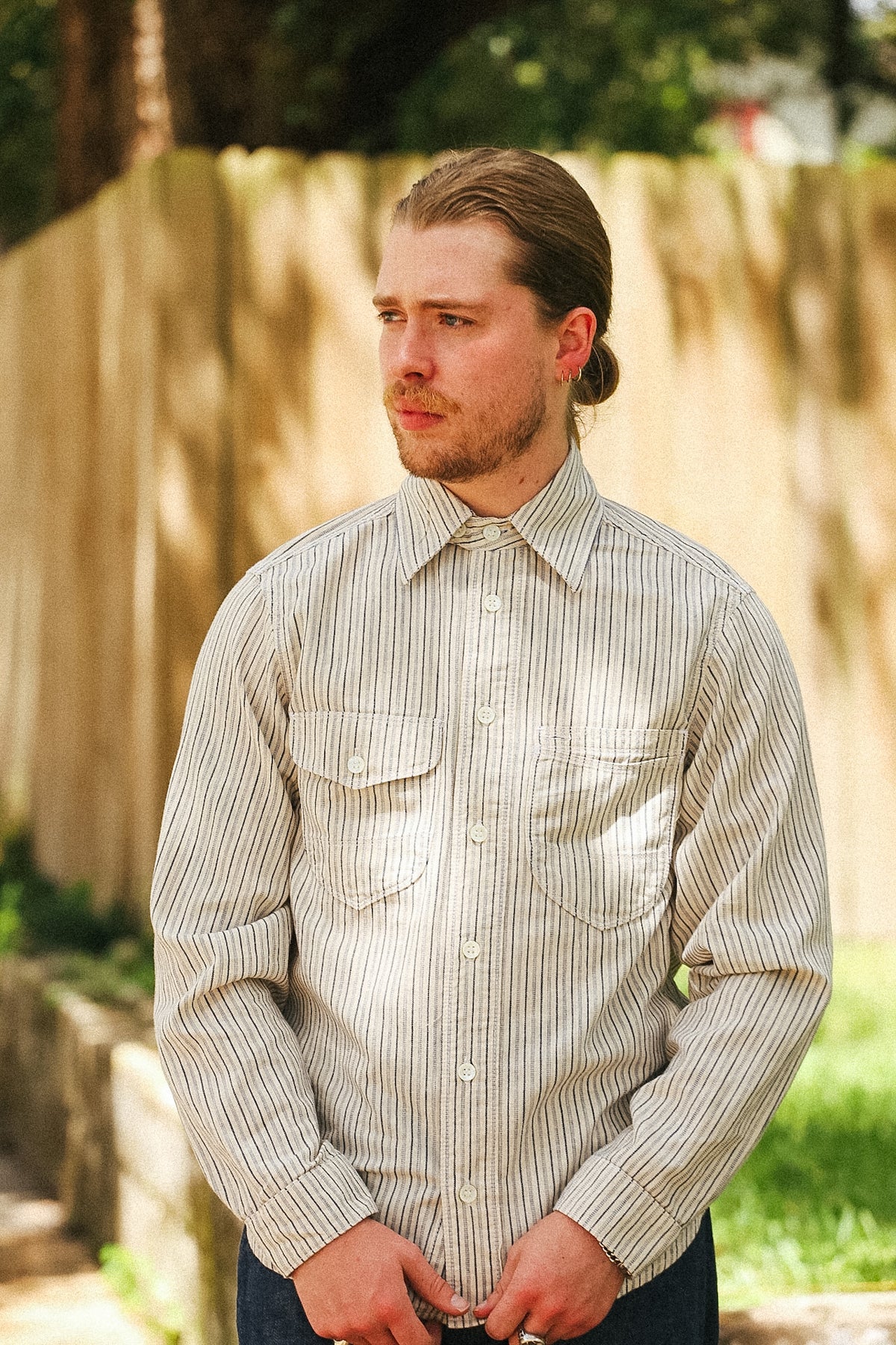 A man wearing a beige and white vertically striped long-sleeve button-down shirt, standing outdoors in front of a wooden fence.