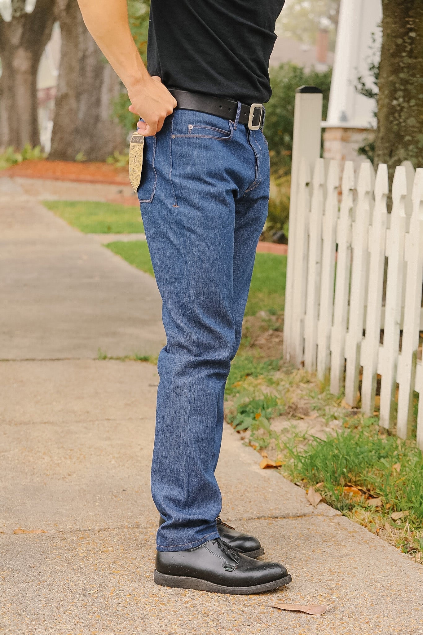 Person wearing blue jeans and black shoes on a sidewalk with a white picket fence in the background.