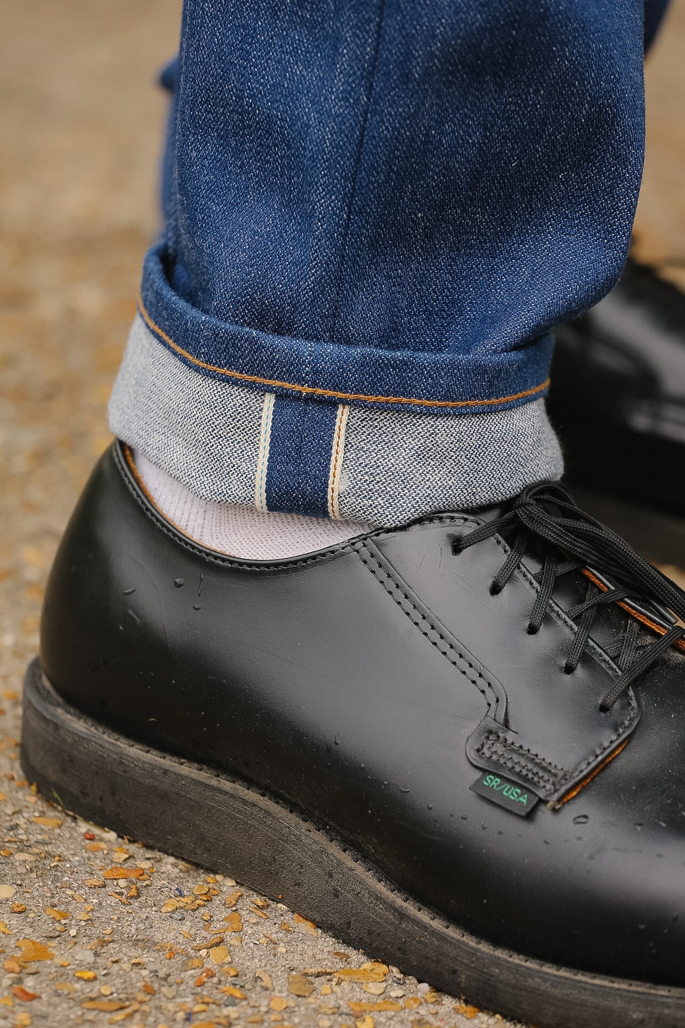 Black leather shoe worn with rolled-up jeans on a textured surface