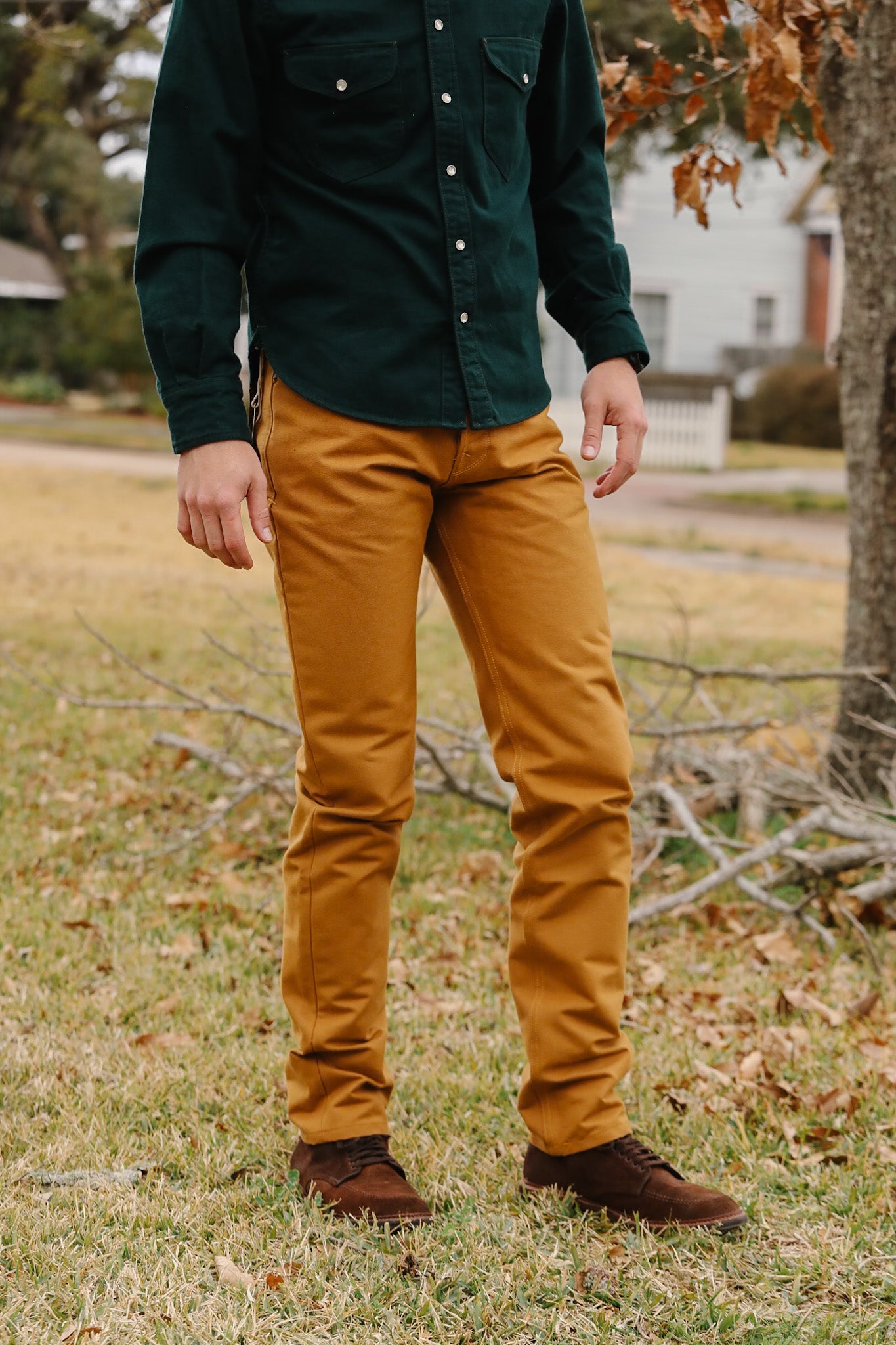 Person wearing a green shirt and mustard yellow pants standing in a grassy area with trees and houses in the background.