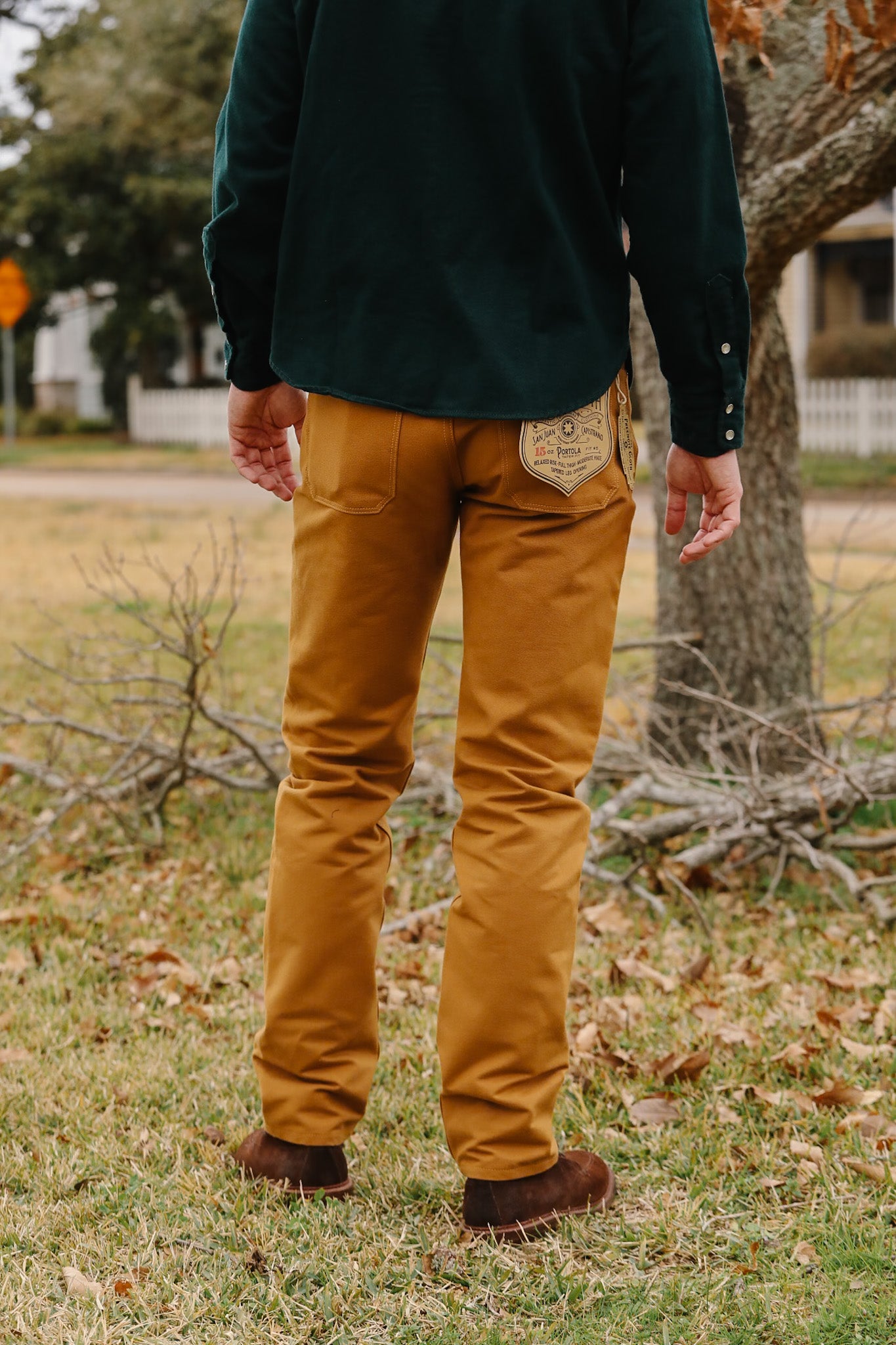 Person wearing mustard pants and a dark jacket standing in a grassy area with trees and a house in the background.