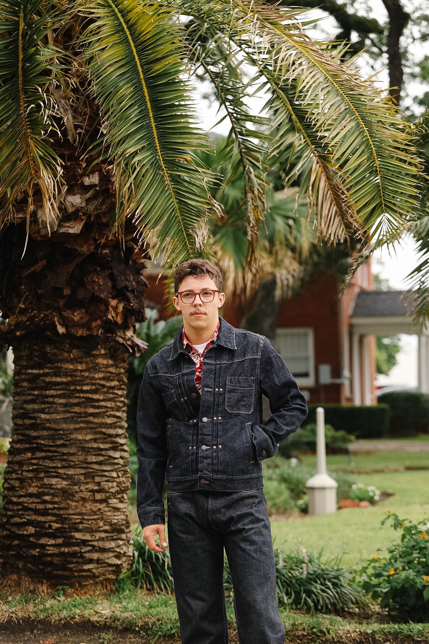 Man standing outdoors under a palm tree with a house in the background