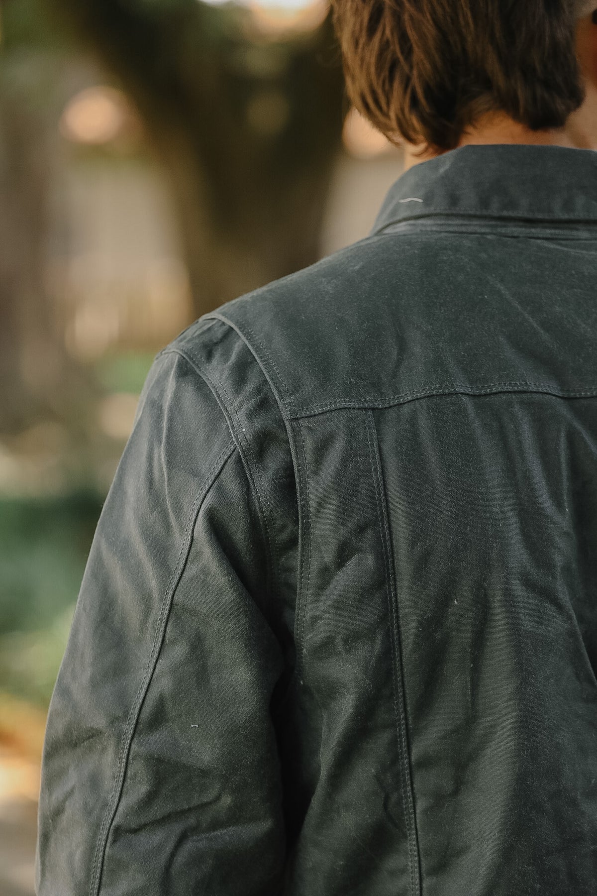 A close-up back view of a person wearing a dark charcoal gray utility jacket in an outdoor setting with a blurred background.