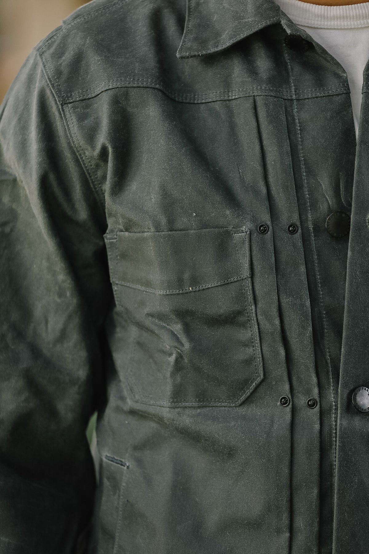 Close-up of a person wearing a dark olive green waxed cotton utility jacket over a white shirt.
