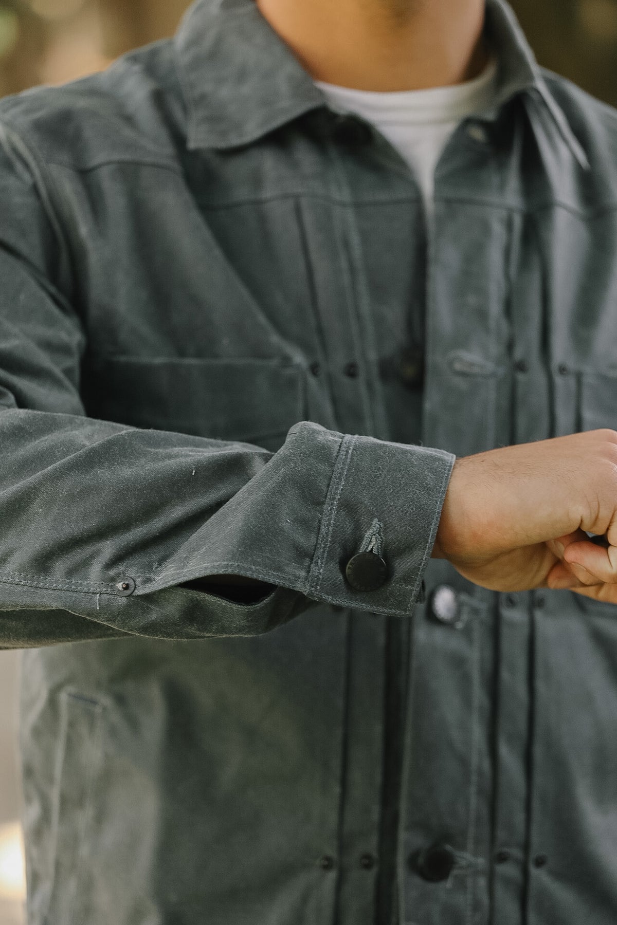 Close-up of a person wearing a dark charcoal gray utility jacket over a white t-shirt, with the right sleeve folded up to show the button cuff.
