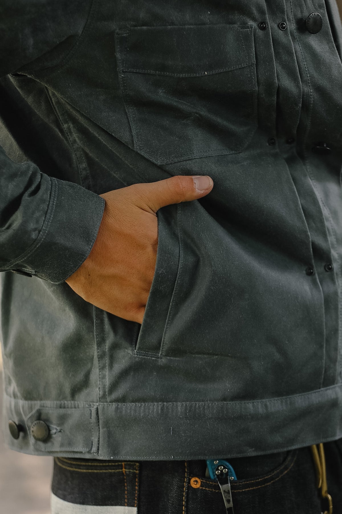 Close-up of a person wearing a dark charcoal gray waxed canvas utility jacket, with a hand tucked into a front pocket.