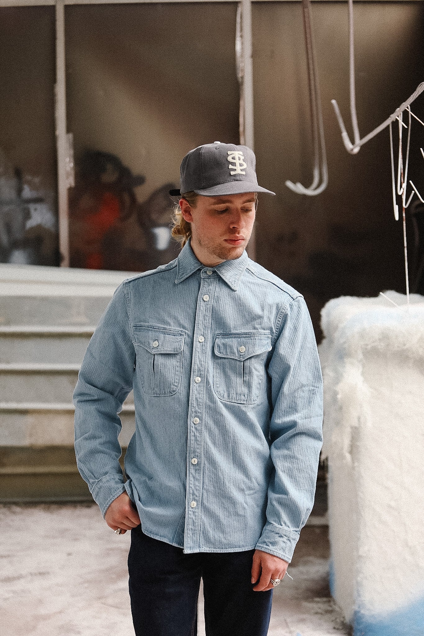 Person wearing a denim shirt and cap standing outdoors with a blurred background
