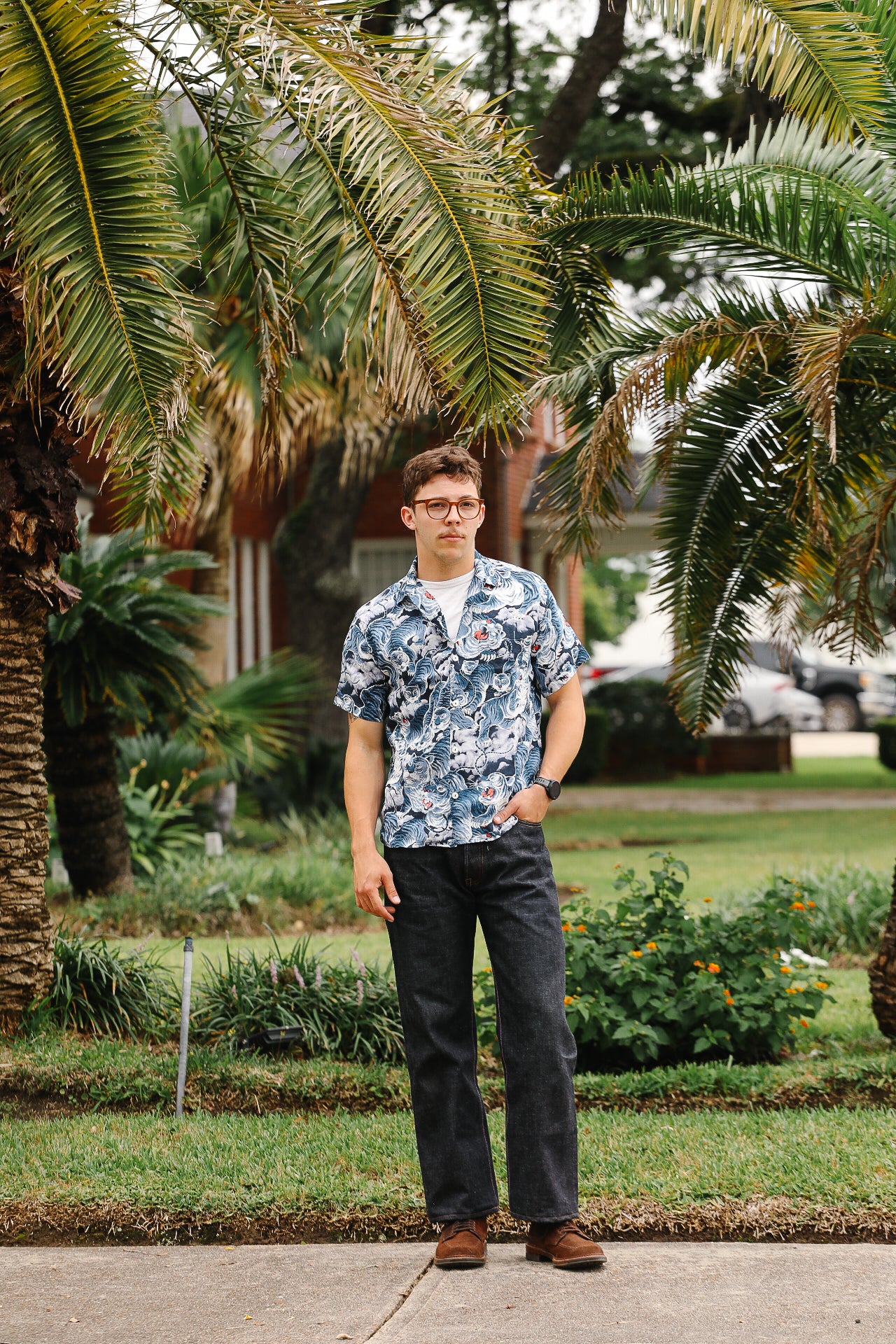 Person wearing a patterned shirt and dark pants standing in a garden with palm trees.