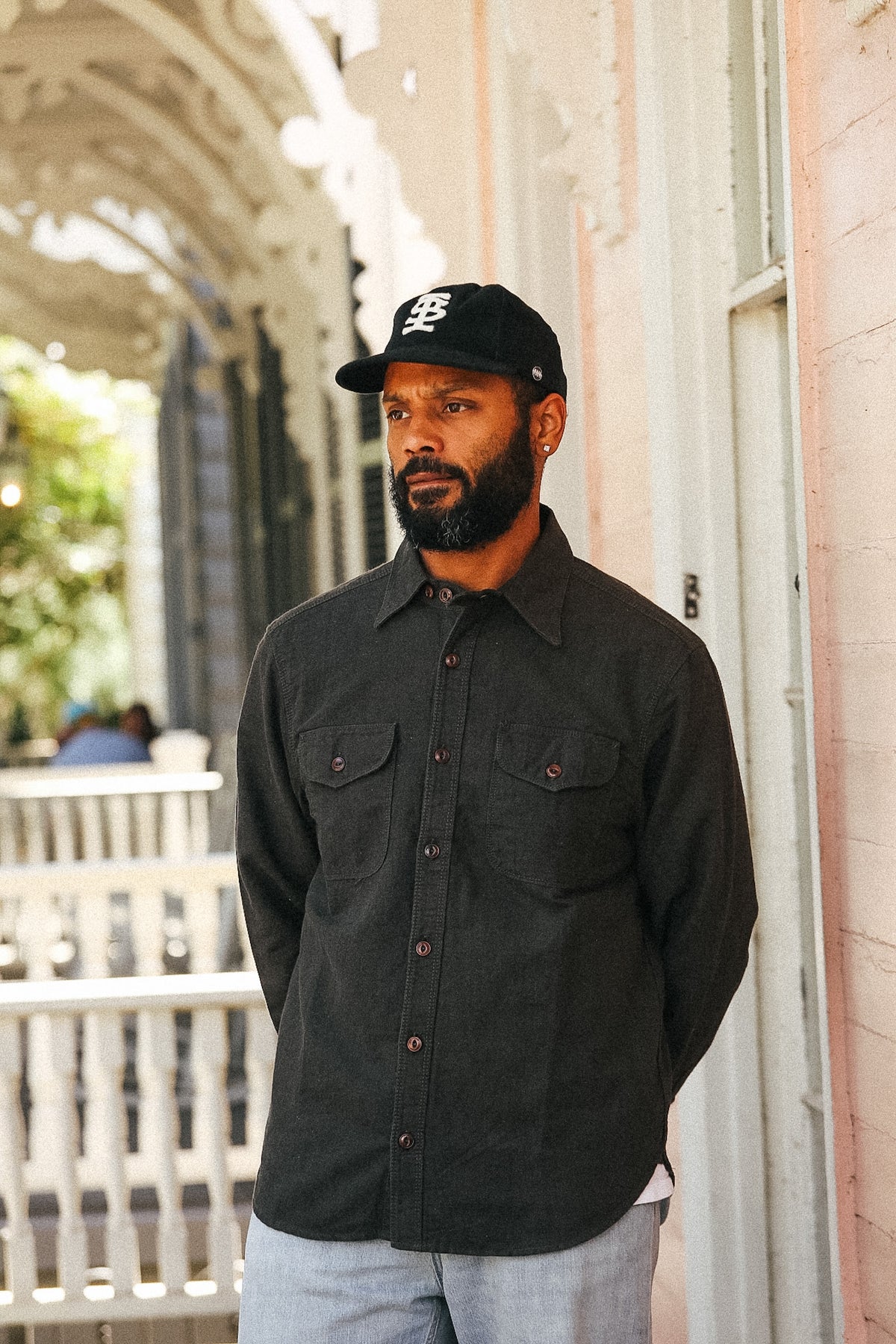 A man wearing a black long-sleeve button-up shirt with chest pockets, a black baseball cap with a white logo, and light gray jeans, standing on a porch next to a pink building.