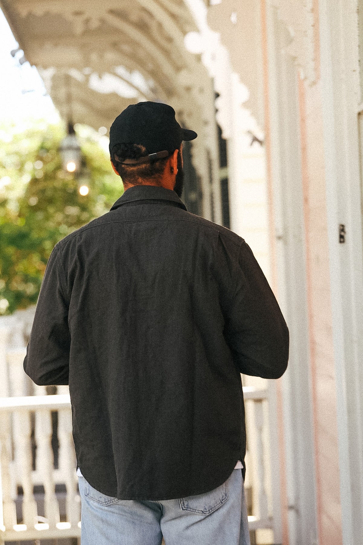 A man viewed from behind wearing a dark charcoal gray long-sleeve button-down shirt and a black baseball cap, standing on a porch.