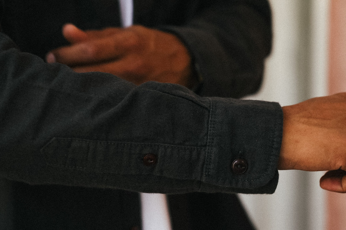 Close-up of a dark charcoal gray button-down shirt sleeve with two buttons, worn by a model with another person's hands visible in the background.