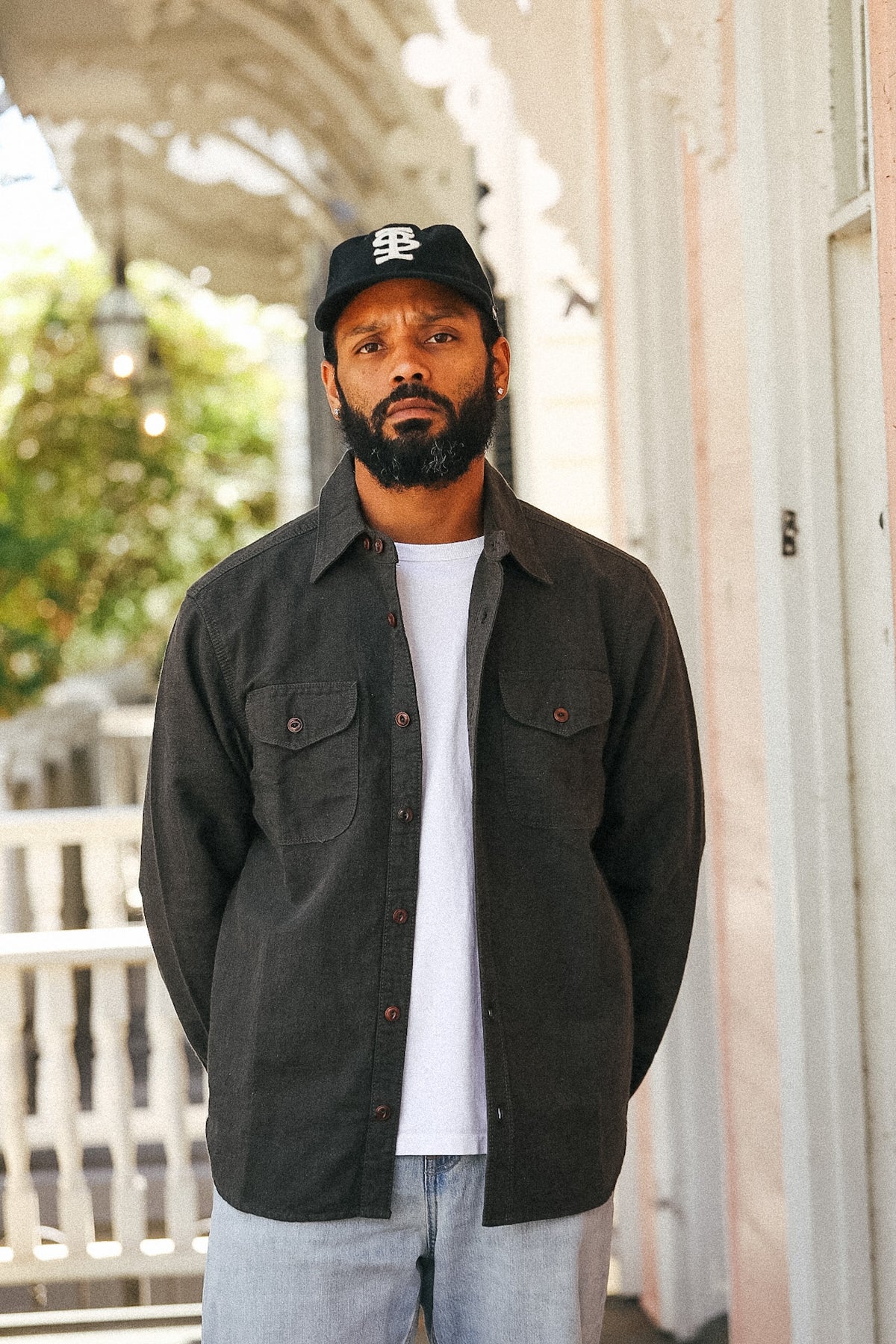 A man wearing a dark charcoal gray button-up overshirt over a white t-shirt, light blue jeans, and a black baseball cap with a white logo, standing on a porch.