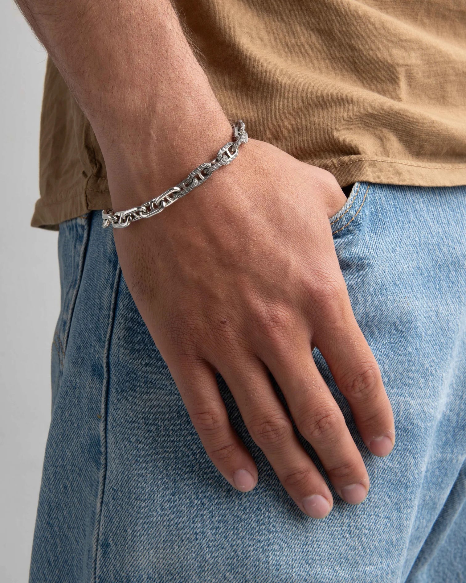 Close-up of a man's hand wearing a sterling silver mixed motif chain bracelet paired with casual jeans and a brown shirt