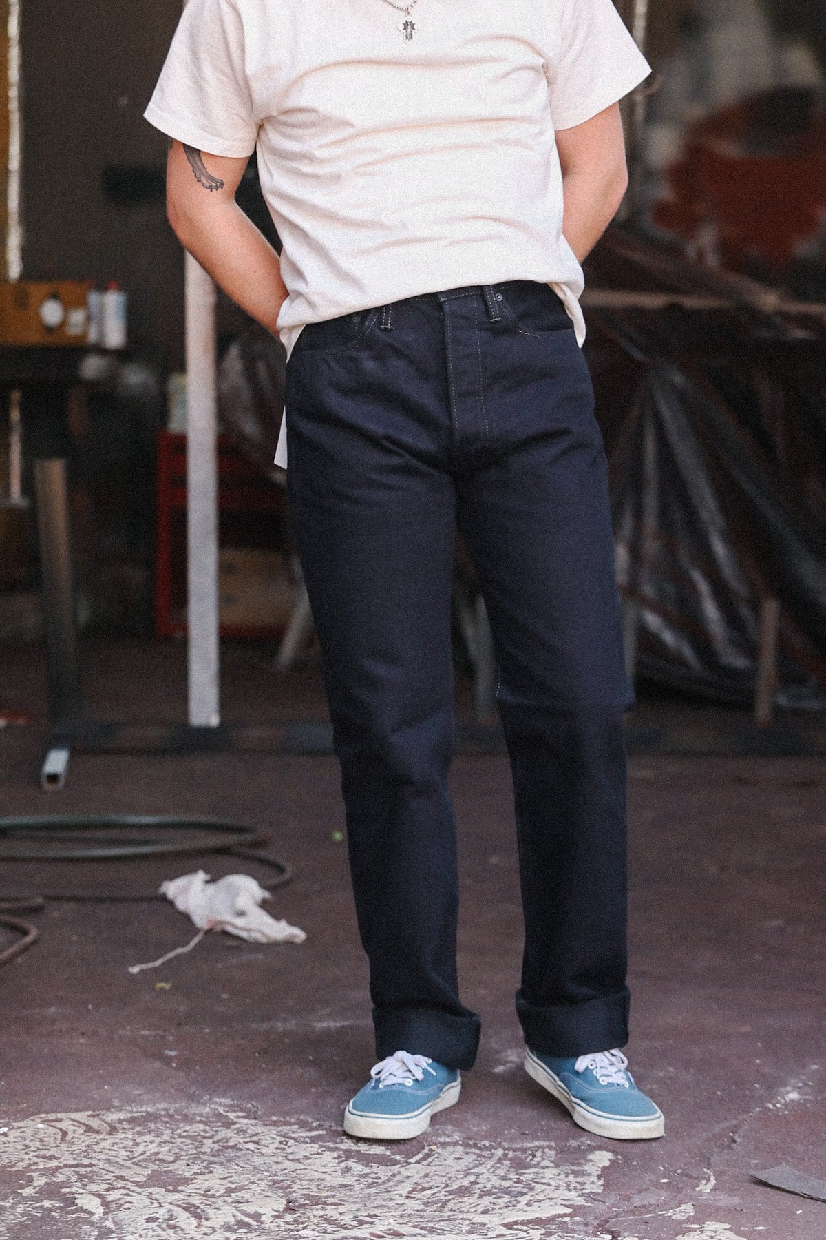 A person wearing dark navy blue straight-leg jeans, a white t-shirt, and light blue sneakers, standing in a workshop environment.