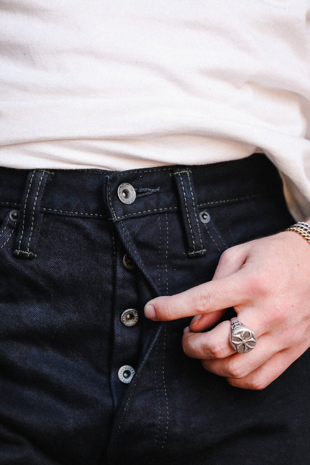 Close-up of a person wearing dark indigo denim jeans with a button fly, a white t-shirt, and a silver ring on their finger.