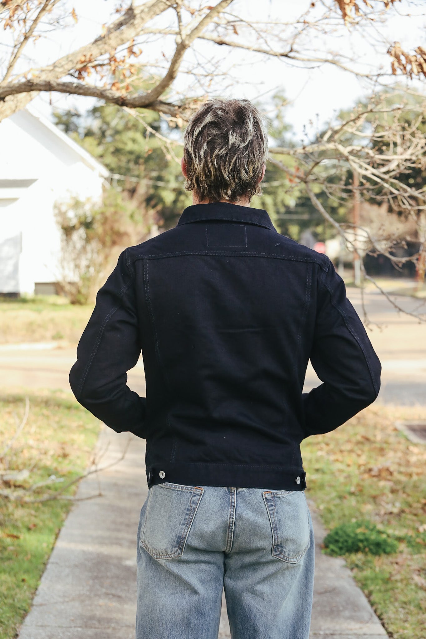 Person wearing a black jacket and light blue jeans standing on a sidewalk with trees and houses in the background.