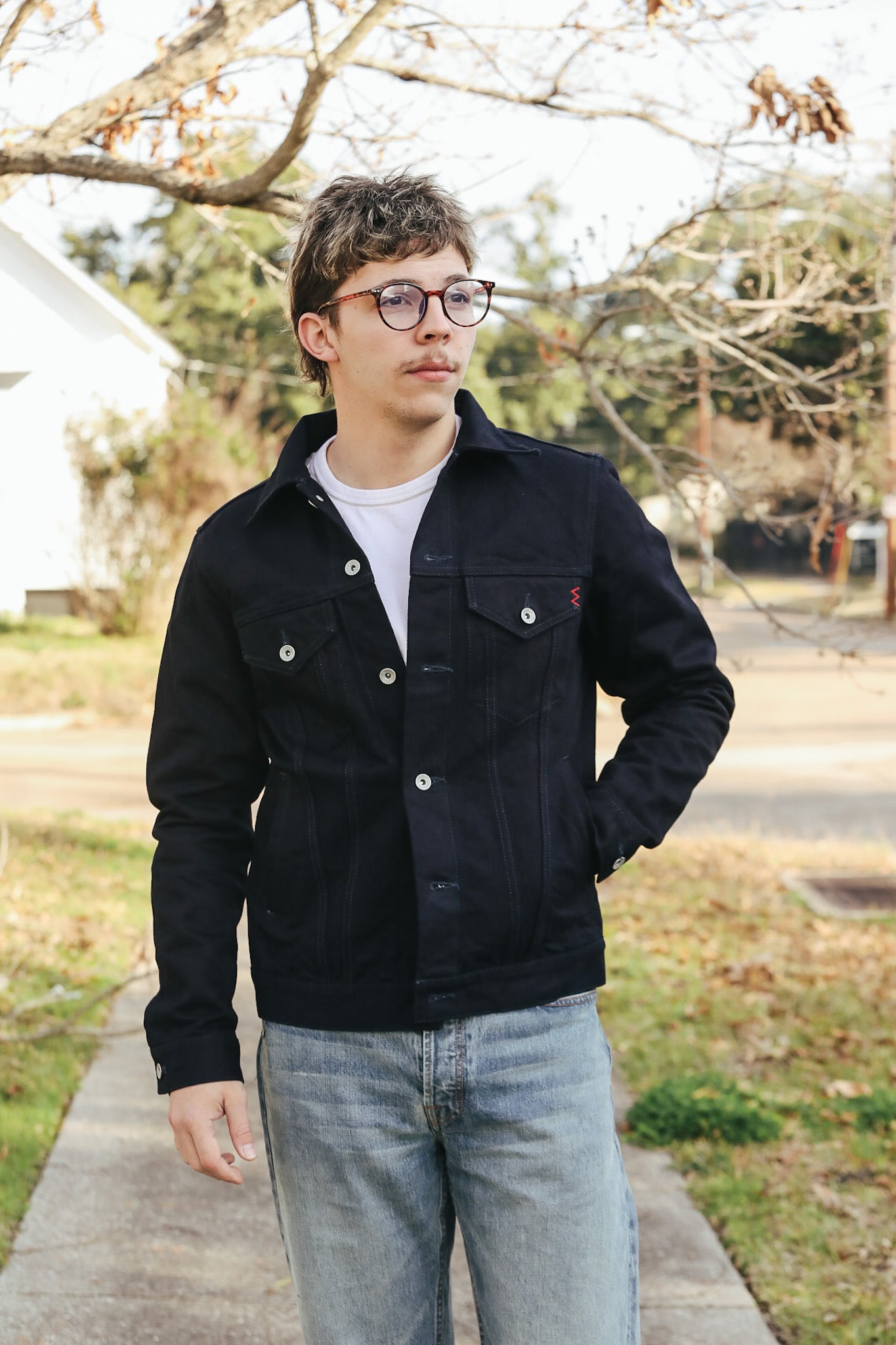 Person wearing a black jacket and glasses standing outdoors on a sidewalk.