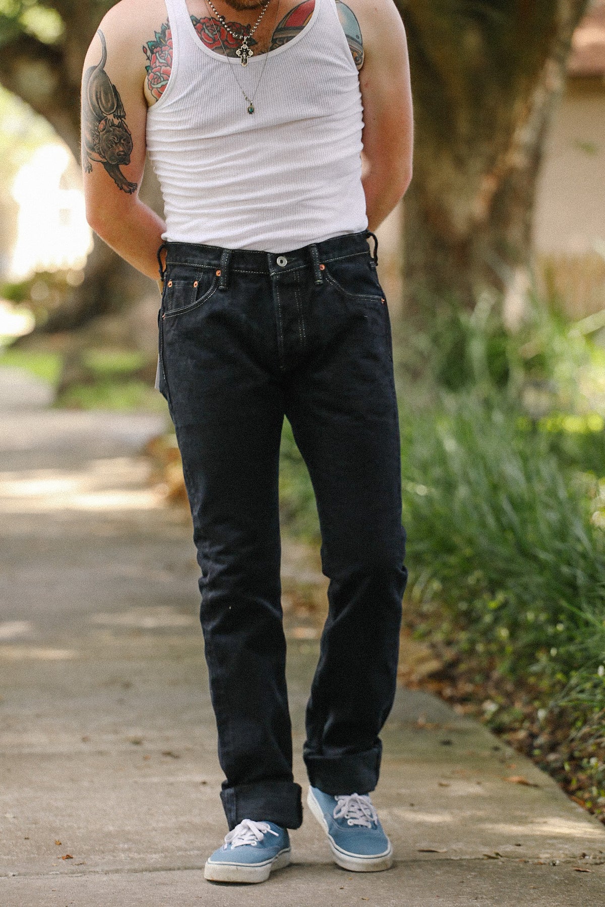 A person standing outdoors on a sidewalk wearing dark blue slim-fit jeans, a white tank top, and light blue sneakers.