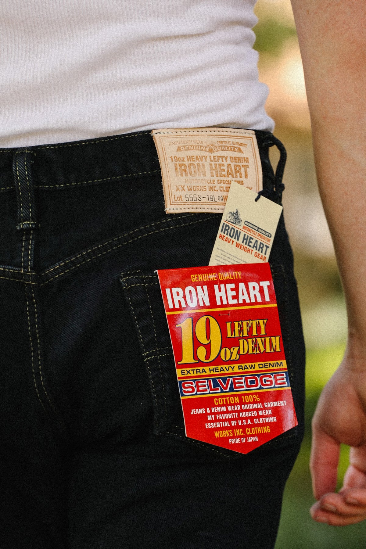 Close-up of the back waistband of black denim jeans with multiple Iron Heart brand tags attached, worn by a person in a white ribbed top against a blurred outdoor background.