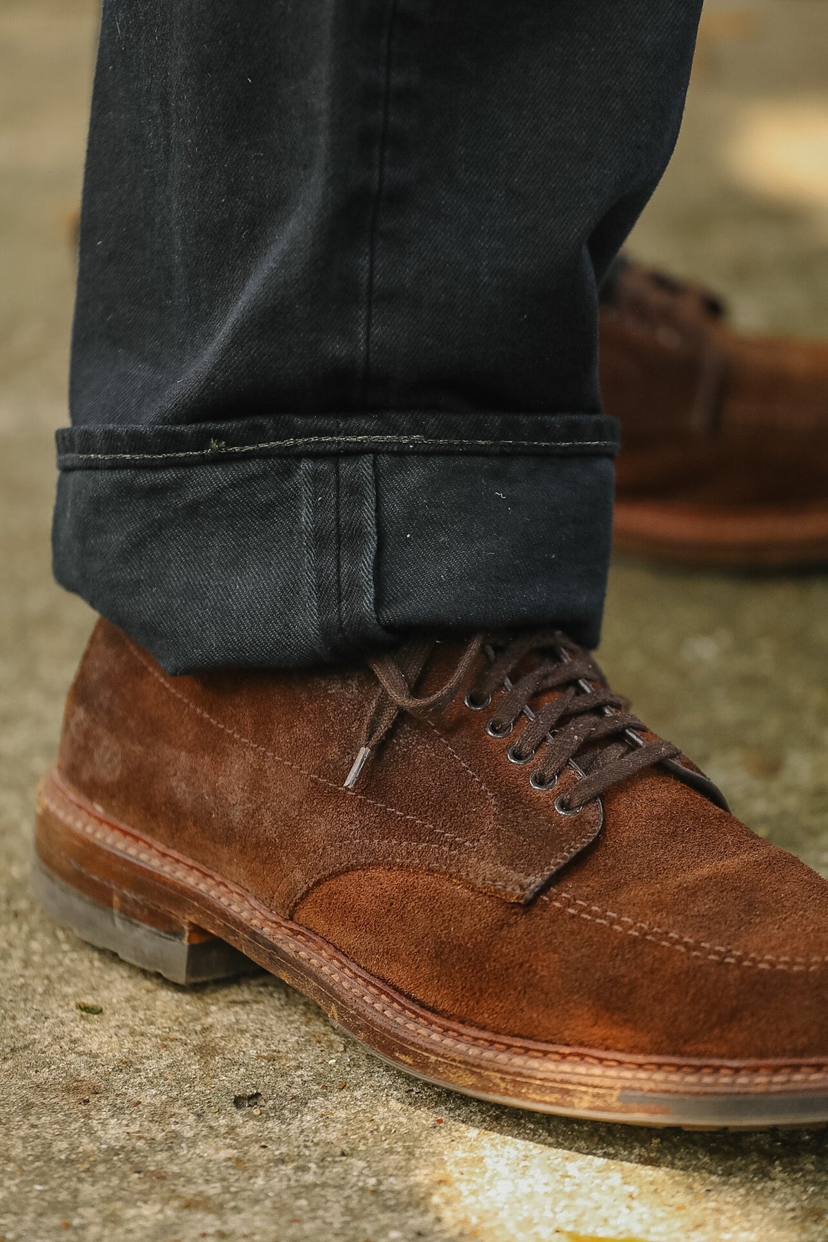 A close-up of a person's foot wearing a brown suede lace-up shoe with dark cuffed jeans, standing on a textured concrete surface.