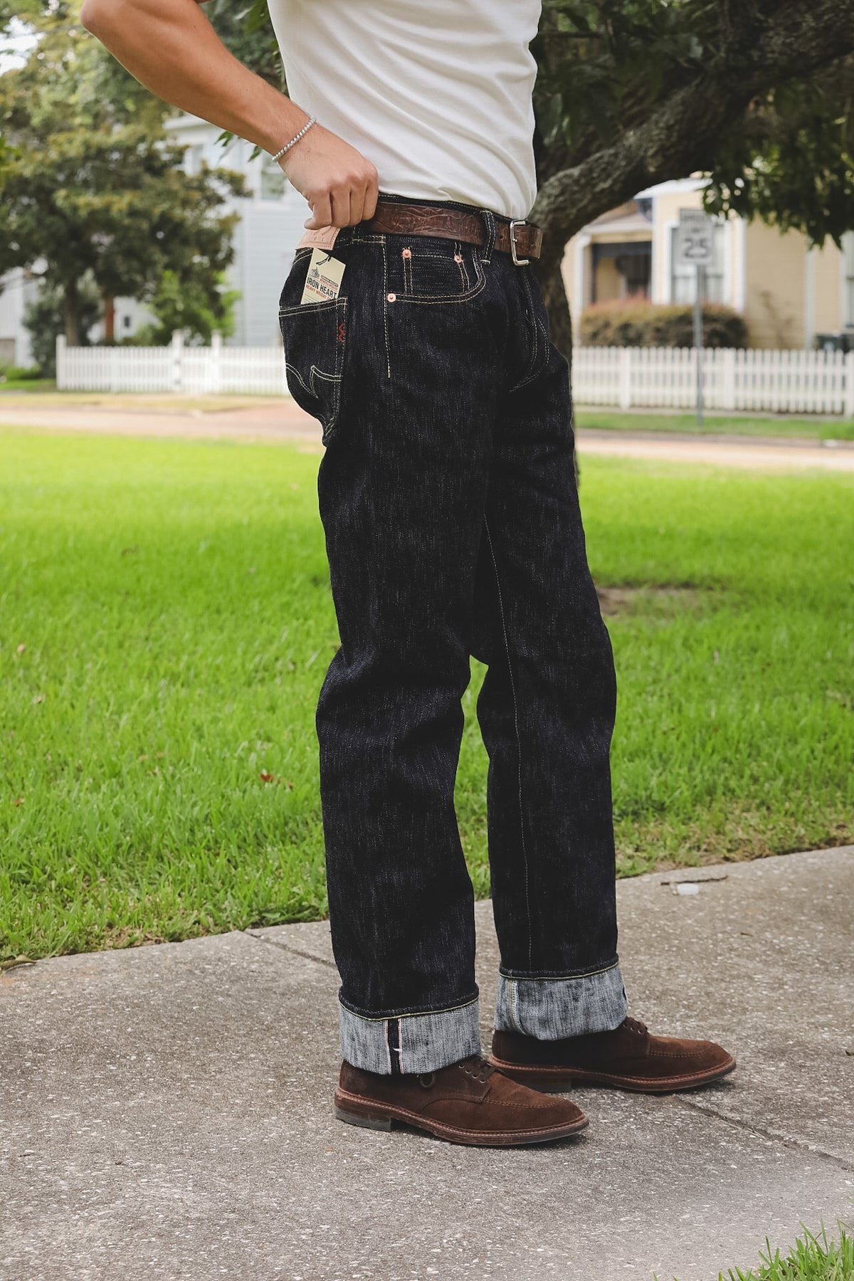 A person outdoors wearing dark indigo denim jeans with a brown belt and brown suede boots, standing on a concrete path with green grass and trees in the background.