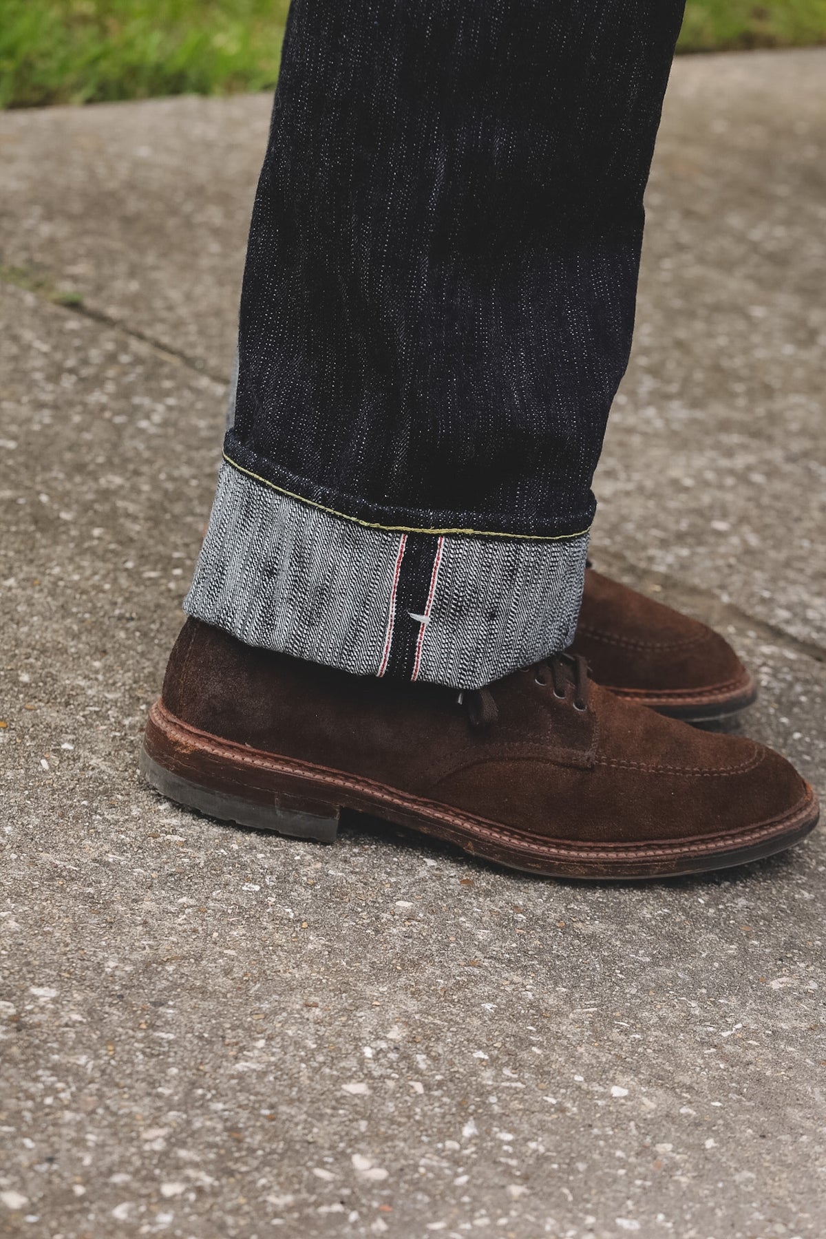 A close-up of a person's legs wearing dark brown suede lace-up shoes and dark blue jeans with the cuffs turned up, standing on a gray concrete surface.