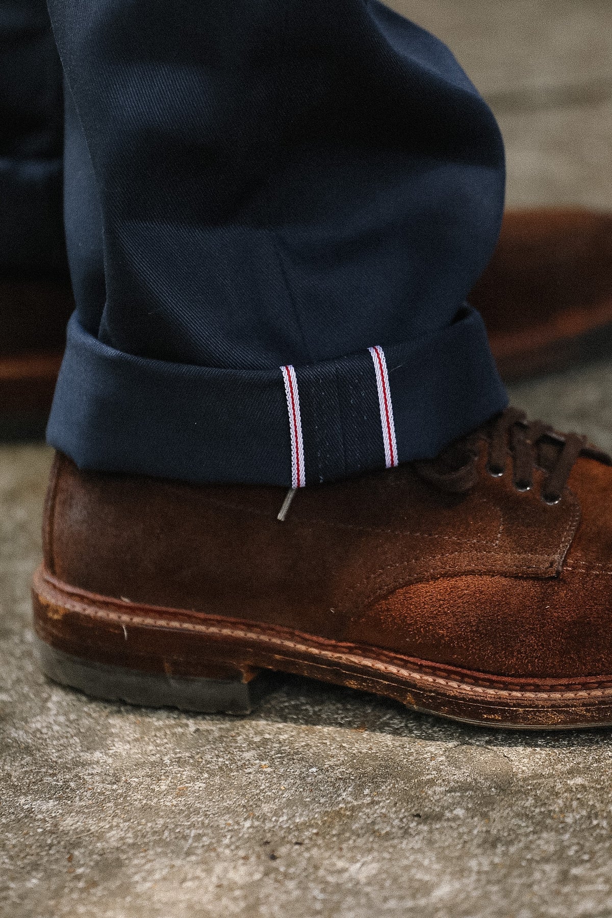 Close-up of a person's ankle wearing navy blue trousers with a rolled cuff showing pink and white stripe detail, paired with a brown suede lace-up shoe on a concrete surface.