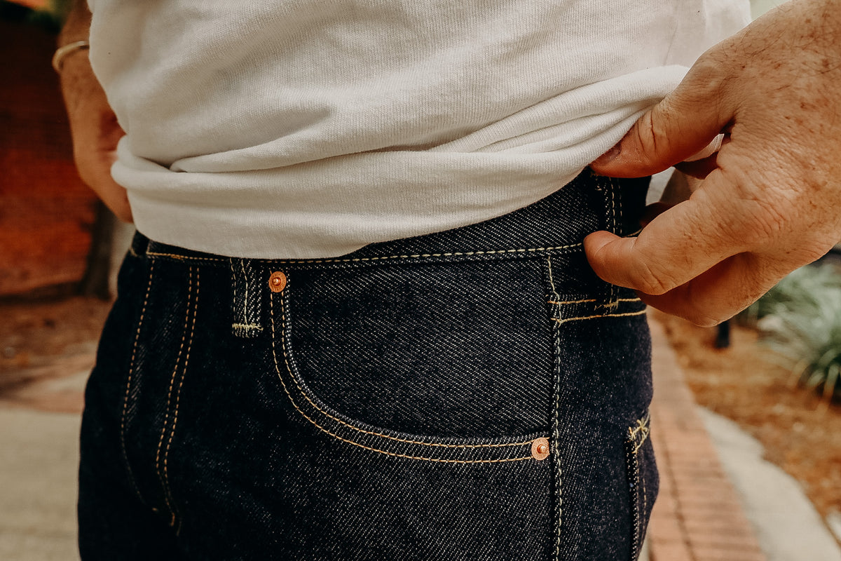A person standing on a sidewalk wearing a white t-shirt and the 21 oz Selvedge Denim Super Slim Tapered Jeans in indigo color with brown shoes.