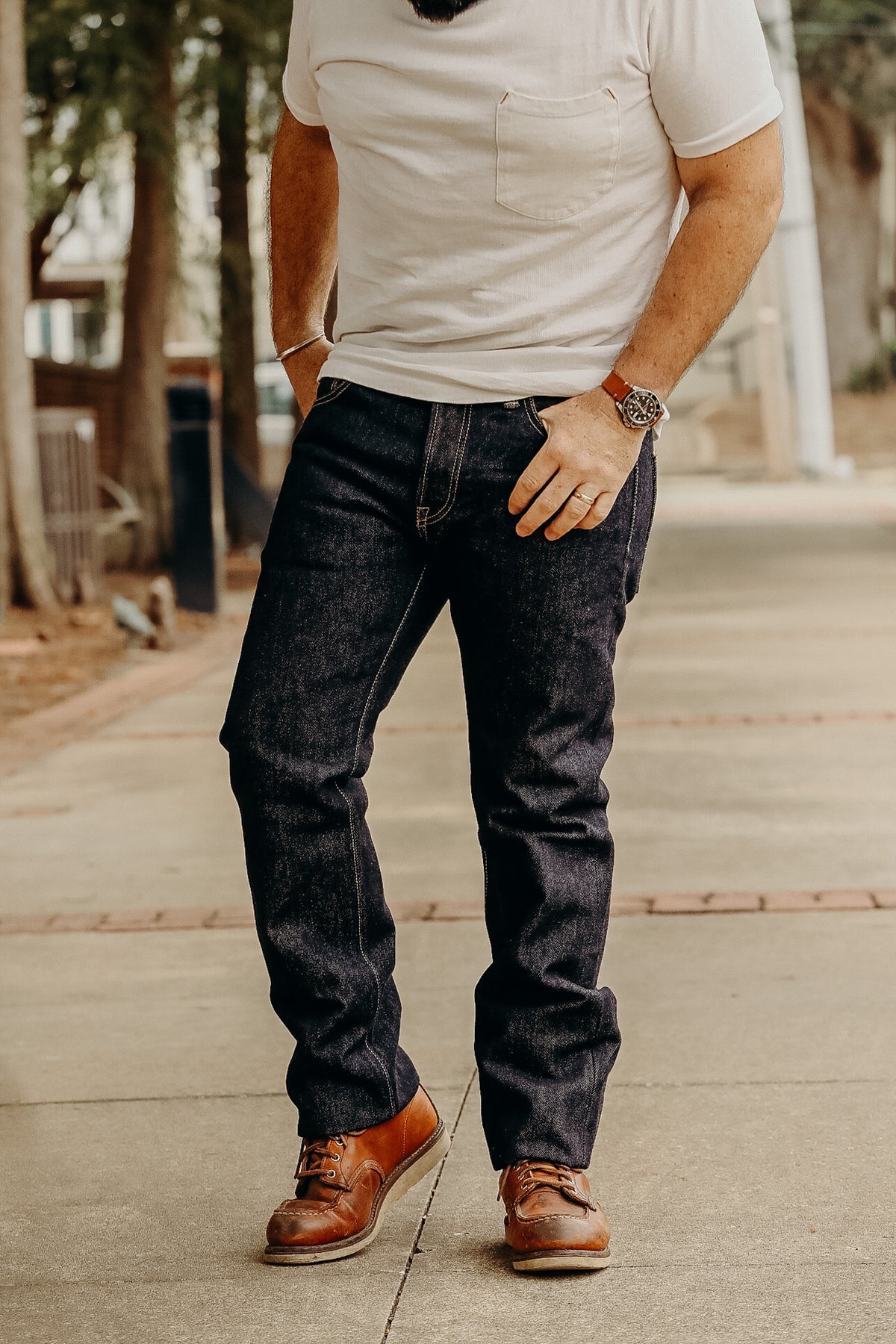 A person standing on a sidewalk wearing a white t-shirt and the 21 oz Selvedge Denim Super Slim Tapered Jeans in indigo color with brown shoes.