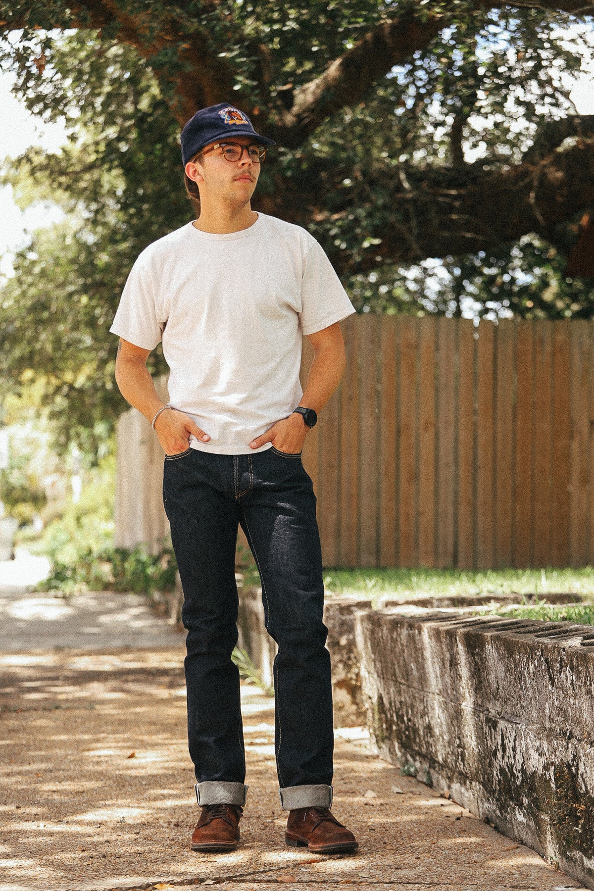 A man standing outdoors on a paved path wearing a white crewneck t-shirt, dark blue jeans, brown boots, and a navy baseball cap with a red logo. Trees and a wooden fence are visible in the background.